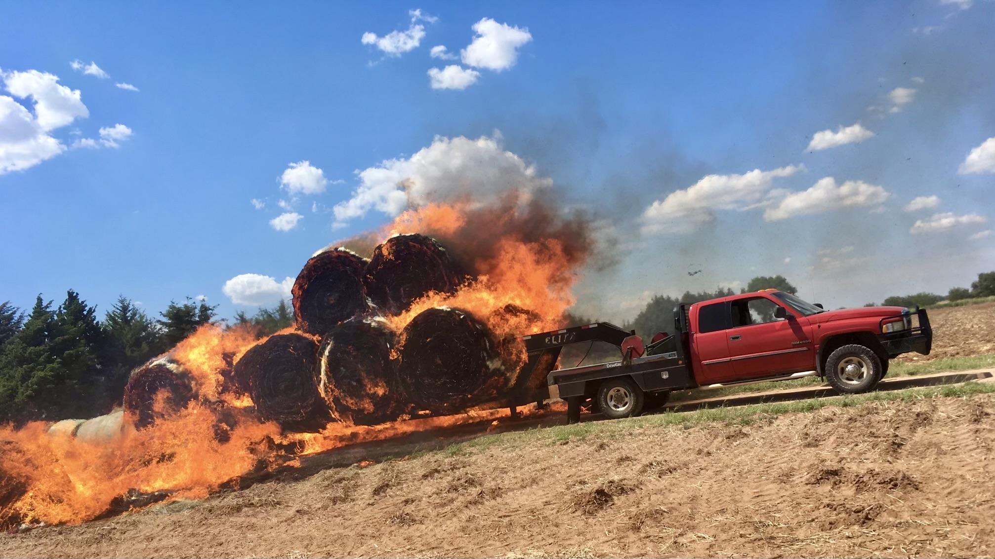 A color, wide-angle photograph in landscape orientation that captures a red heavy-duty pickup truck pulling a flatbed gooseneck trailer loaded with large round hay bales that have erupted into a massive, intense fire. The fire consumes the entire load, with bright orange and yellow flames licking high into the air and thick, dark grey smoke billowing toward a bright blue sky dotted with fluffy white clouds. Several charred bales have already fallen off the back of the trailer onto the field, continuing to burn and creating a trail of fire behind the vehicle. The red truck, equipped with a black flatbed and a front brush guard, is stopped on a harvested field. The surrounding landscape consists of dry, brown earth and a line of dark green evergreen trees in the distance. The lighting is bright and clear, highlighting the sharp contrast between the vibrant red of the truck and the destructive glow of the hay fire.