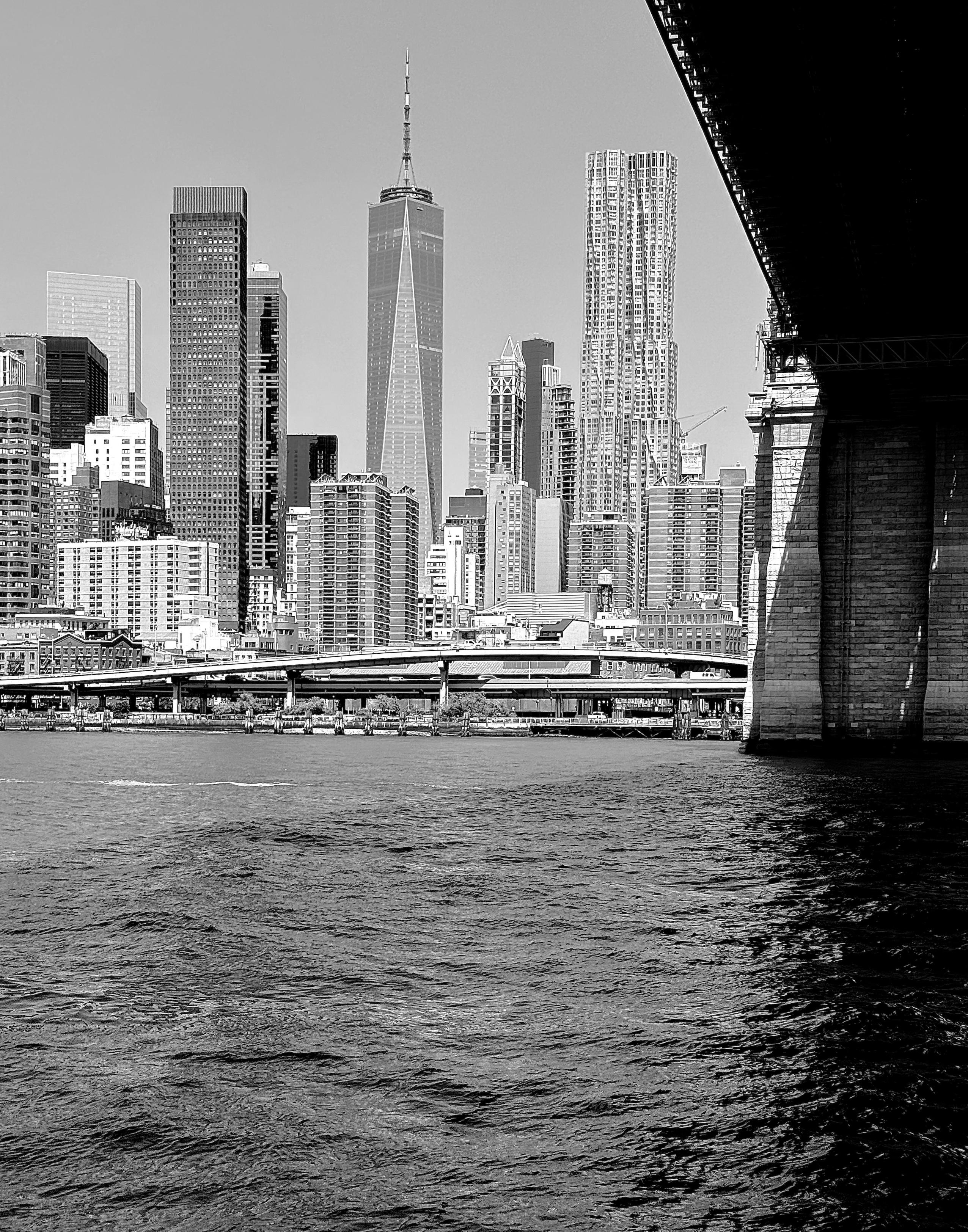A noir, wide-angle photograph that captures the dense skyline of Lower Manhattan as seen from atop a ferry in the East River. Taken from a low vantage point, the composition is framed on the right by the massive, weathered stone anchorage and dark underside of the Brooklyn Bridge. The choppy water in the foreground is rendered in deep, high-contrast tones, leading the eye toward the FDR Drive elevated highway that hugs the shoreline. The midday light creates sharp shadows and emphasizes the architectural textures of the city, ranging from the smooth glass of modern skyscrapers to the rhythmic patterns of older apartment blocks. Dominating the center-left of the frame is the soaring One World Trade Center, its geometric facets catching the light against a clear, pale sky. To its right stands the rippling, metallic facade of the Gehry Building, which provides a stark, liquid-like contrast to the rigid rectangles of the surrounding structures. The monochrome palette strips away the city’s usual color, focusing instead on the dramatic play between light and shadow, the immense scale of the suspension bridge, and the sheer verticality of the urban landscape.