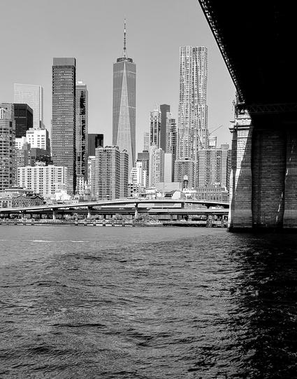 A noir, wide-angle photograph that captures the dense skyline of Lower Manhattan as seen from atop a ferry in the East River. Taken from a low vantage point, the composition is framed on the right by the massive, weathered stone anchorage and dark underside of the Brooklyn Bridge. The choppy water in the foreground is rendered in deep, high-contrast tones, leading the eye toward the FDR Drive elevated highway that hugs the shoreline. The midday light creates sharp shadows and emphasizes the architectural textures of the city, ranging from the smooth glass of modern skyscrapers to the rhythmic patterns of older apartment blocks. Dominating the center-left of the frame is the soaring One World Trade Center, its geometric facets catching the light against a clear, pale sky. To its right stands the rippling, metallic facade of the Gehry Building, which provides a stark, liquid-like contrast to the rigid rectangles of the surrounding structures. The monochrome palette strips away the city’s usual color, focusing instead on the dramatic play between light and shadow, the immense scale of the suspension bridge, and the sheer verticality of the urban landscape.