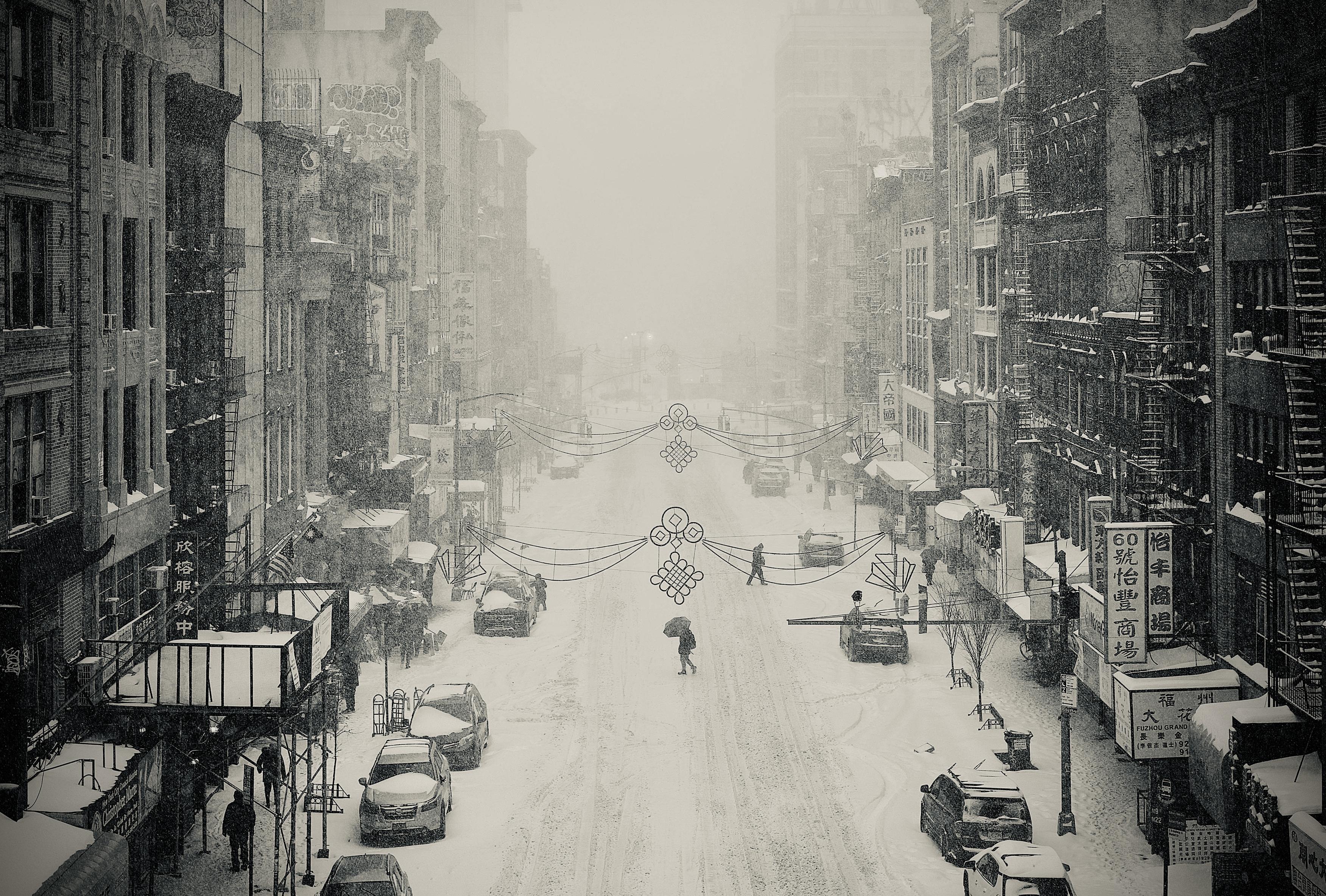 A silver tone, wide-angle photograph in landscape orientation that captures an atmospheric, high-angle scene above Broadway in the Chinatown neighborhood of Manhattan during a heavy snowstorm. The perspective looks down the center of the street, where a thick blanket of white snow covers the pavement and parked cars, showing only faint tire tracks. In the center of the frame, a lone figure with a dark umbrella crosses the street, emphasizing a sense of quiet isolation. Ornate, traditional Chinese decorative banners are strung across the air between buildings, their intricate patterns standing out against the dense, foggy white background where the street eventually disappears into the haze. The flanking architecture consists of tall, weathered urban buildings with visible fire escapes and storefronts adorned with Chinese signage. The left side features scaffolding and a few pedestrians huddled near the building edges, while the right side shows parked vehicles almost completely overtaken by snow. The lighting is soft and diffused, typical of a blizzard, creating a dreamlike, cinematic quality that blurs the line between a modern city and a historical memory. The overall composition is balanced and serene, highlighting the stillness that a major snowstorm brings to an otherwise bustling neighborhood.