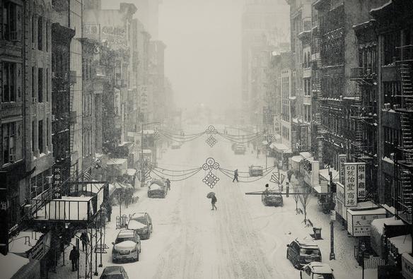 A silver tone, wide-angle photograph in landscape orientation that captures an atmospheric, high-angle scene above Broadway in the Chinatown neighborhood of Manhattan during a heavy snowstorm. The perspective looks down the center of the street, where a thick blanket of white snow covers the pavement and parked cars, showing only faint tire tracks. In the center of the frame, a lone figure with a dark umbrella crosses the street, emphasizing a sense of quiet isolation. Ornate, traditional Chinese decorative banners are strung across the air between buildings, their intricate patterns standing out against the dense, foggy white background where the street eventually disappears into the haze. The flanking architecture consists of tall, weathered urban buildings with visible fire escapes and storefronts adorned with Chinese signage. The left side features scaffolding and a few pedestrians huddled near the building edges, while the right side shows parked vehicles almost completely overtaken by snow. The lighting is soft and diffused, typical of a blizzard, creating a dreamlike, cinematic quality that blurs the line between a modern city and a historical memory. The overall composition is balanced and serene, highlighting the stillness that a major snowstorm brings to an otherwise bustling neighborhood.