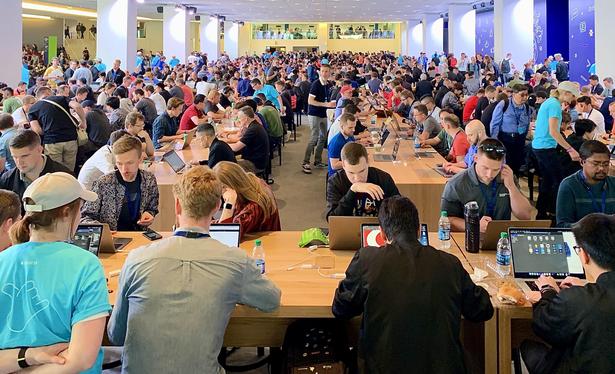 A color, wide-angle photograph in landscape orientation that captures a massive, brightly lit hall within the McEnery Convention Center, packed with hundreds of attendees following an Apple Worldwide Developers Conference (WWDC) keynote in 2019. Rows of long, light-colored wooden communal tables stretch deep into the background, almost entirely occupied by developers focused on their open laptops. Many individuals are leaning forward or resting their heads in their hands as they wait for new beta software releases to download. A sea of people in casual attire fills the space, creating a dense mosaic of tech enthusiasts engaged in the same post-keynote ritual. In the foreground, the focus is on several developers seated at a table, their screens displaying various coding environments and system interfaces. One individual on the left wears a bright cyan WWDC19 t-shirt, while others have scattered snacks, water bottles, and tangled charging cables across the wooden surface. The architecture of the hall features large white square pillars and a distant mezzanine level where more crowds are visible, adding to the sense of scale. The lighting is soft and even, reflecting off the floor and the numerous aluminum laptop lids.