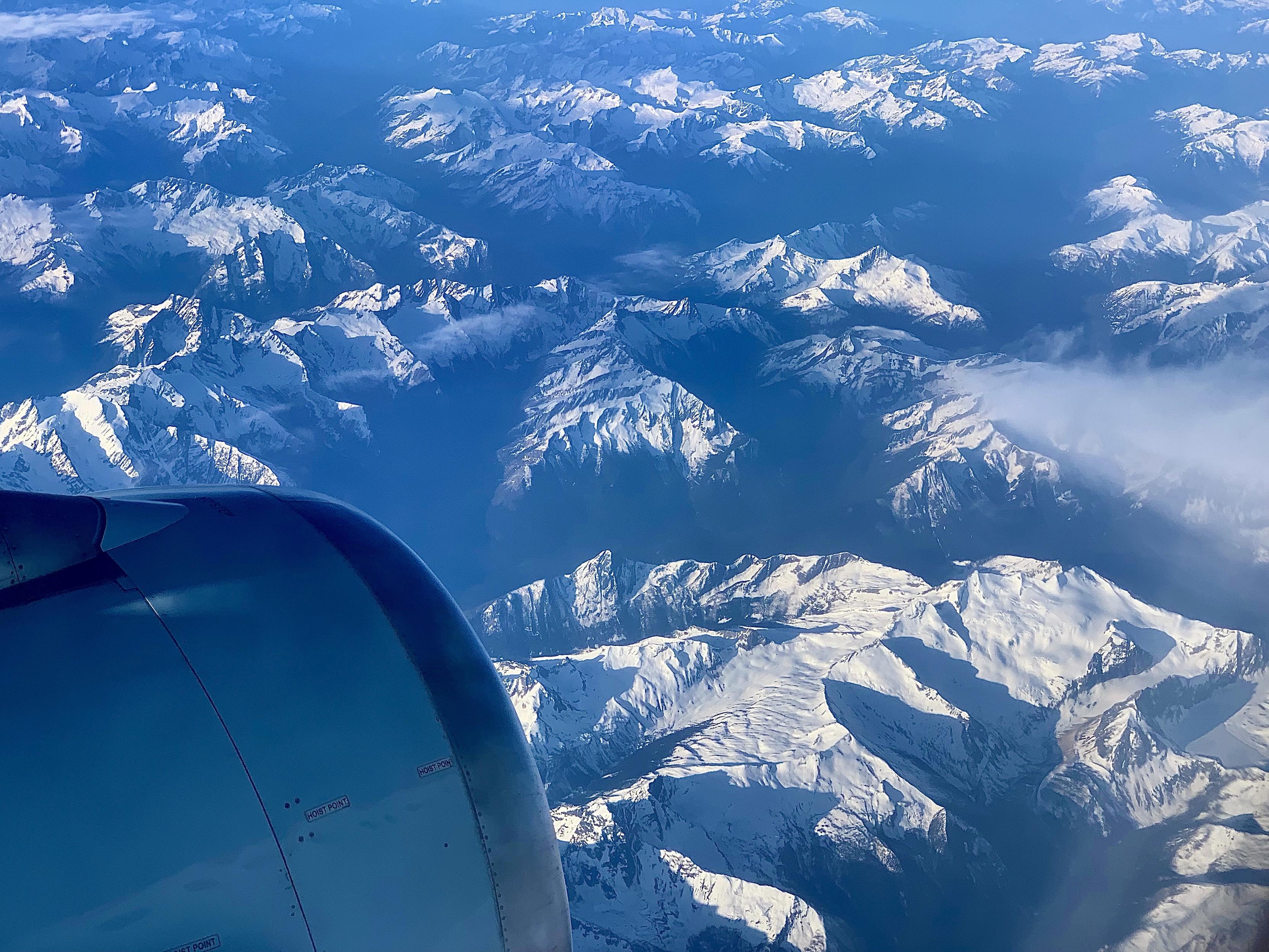 A color, wide-angle photograph captured from a high-altitude passenger jet window looking down upon a vast expanse of the Rocky Mountains in Colorado. The foreground is dominated by the smooth, curved metallic surface of a jet engine intake, its blue-grey casing reflecting the bright sky. Below and beyond the aircraft, a sea of jagged, snow-capped peaks and deep, shadowed valleys stretches toward the horizon. The terrain is a stark contrast of bright white snow clinging to the ridges and the dark, textured rock of the steep vertical faces. The lighting shows a clear, bright day, casting deep, cool-toned blue shadows across the eastern slopes and valley floors, which emphasizes the extreme verticality of the mountain range. Thin, wispy stratus clouds drift between some of the lower peaks, partially obscuring the depths of the canyons.