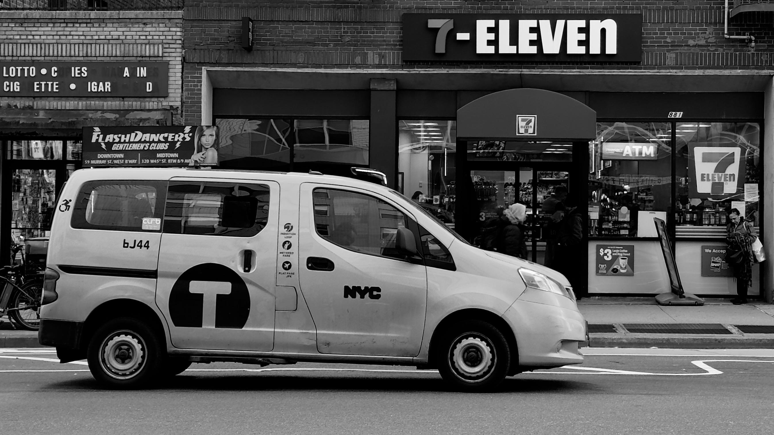 A noir, medium-angle photograph in landscape orientation that captures the street scene along Eighth Avenue in the Hell’s Kitchen neighborhood of Manhattan. The focal point of the image is a prominent NYC Taxi van in the foreground. The vehicle is oriented to the right and displays a large "T" logo inside a circle on its side door, alongside "NYC" lettering and the taxi number "6J44." Small icons indicating accessibility features and payment options are visible near the rear window. The van has a small advertisement board installed above the back window for "FlashDancers Gentlemen’s Clubs." The van is driving slowly towards the right edge of the frame. In the background, the storefront of a 7-Eleven convenience store dominates the upper right quadrant. The store features a dark facade with a large, backlit "7-ELEVEN" sign above a red awning. Through the glass windows, customers and shelves of merchandise are visible, and a sidewalk ATM sign is placed near the entrance. To the left of the 7-Eleven, a more weathered building displays a vintage-style marquee with partial lettering for "LOTTO," "CIGARETTES," and "CIGARS.” A few pedestrians are blurred in the background, including one person standing near the store entrance panhandling. An overcast winter afternoon sky is evening illuminating the scene.