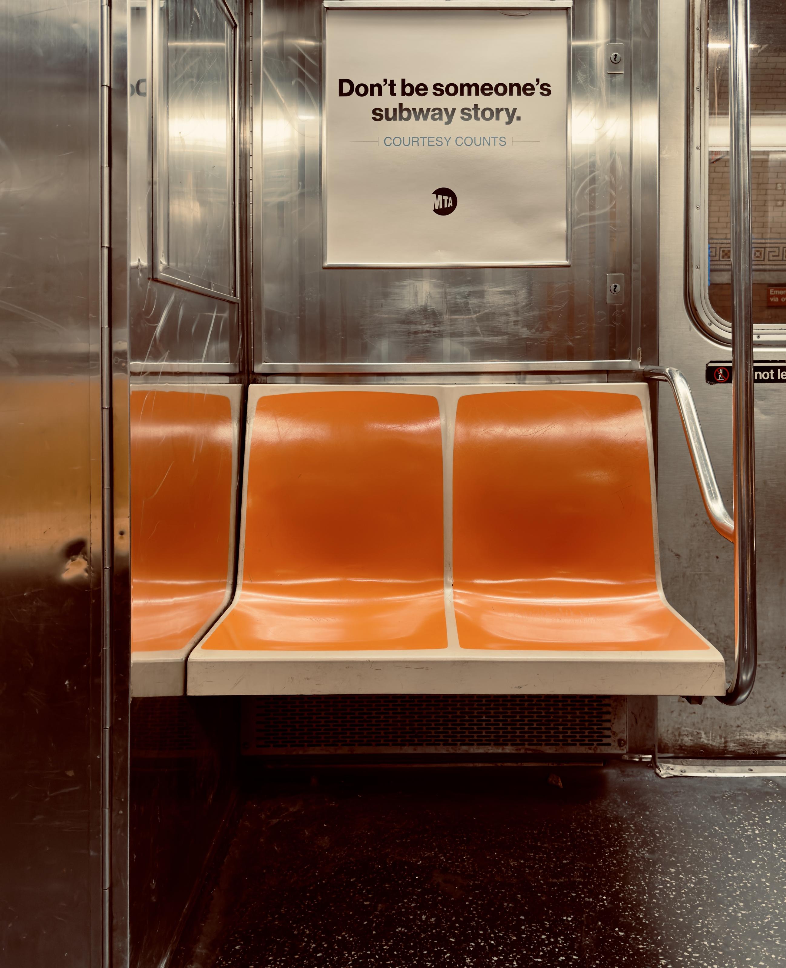 A color, medium-angle photograph in portrait orientation that captures a quiet, symmetrical interior view of a New York City 1 train subway car. In the center, two empty, molded plastic seats are joined together, featuring a vibrant, slightly weathered orange color that pops against the industrial surroundings. Above the seats, an MTA public service announcement poster is mounted on the brushed stainless steel wall. The poster features a clean, minimalist design with the bold text, "Don’t be someone’s subway story," followed by the smaller tagline "Courtesy Counts" and the circular MTA logo. The overall aesthetic is metallic and utilitarian, dominated by reflective stainless steel panels that show signs of minor scratches and wear from daily use. To the left, a vertical metal partition reflects the orange of the seats, while to the right, a curved silver handrail provides a sense of depth. To the right, the platform of 181st Street Station is partially visible through a window on the train car door. The floor is a dark, speckled gray material typical of older transit cars, and the lighting is warm yet industrial, casting soft shadows beneath the seats and highlighting the textures of the subway’s interior.