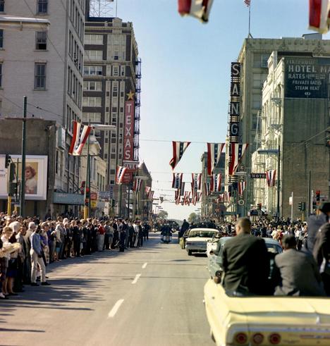 A color, medium-angle, historic photograph that captures Main Street in Dallas during the presidential motorcade on November 22, 1963. The scene is densely packed with spectators lining both sides of the wide, sun-drenched asphalt road. Red, white, and blue bunting and American flags are strung across the street overhead. In the distance, a group of police motorcycles leads the procession away from the camera, followed by several mid-century sedans. In the immediate foreground, the back of a pale yellow convertible is visible, with two men in suits seated in the rear, their backs to the viewer. The architectural landscape features several prominent mid-20th-century buildings and vintage signage. On the left, a tall brick building displays a vertical neon sign for the "Texas Bank," topped with a yellow star. On the right, signs for the "Hotel Mayfair" and the "Sanger Hotel" are visible against the clear blue sky. The sidewalks are a sea of people in period-accurate attire—men in fedoras and suits, and women in dresses—all leaning forward to catch a glimpse of the motorcade. The lighting is bright and sharp, casting long shadows across the street.