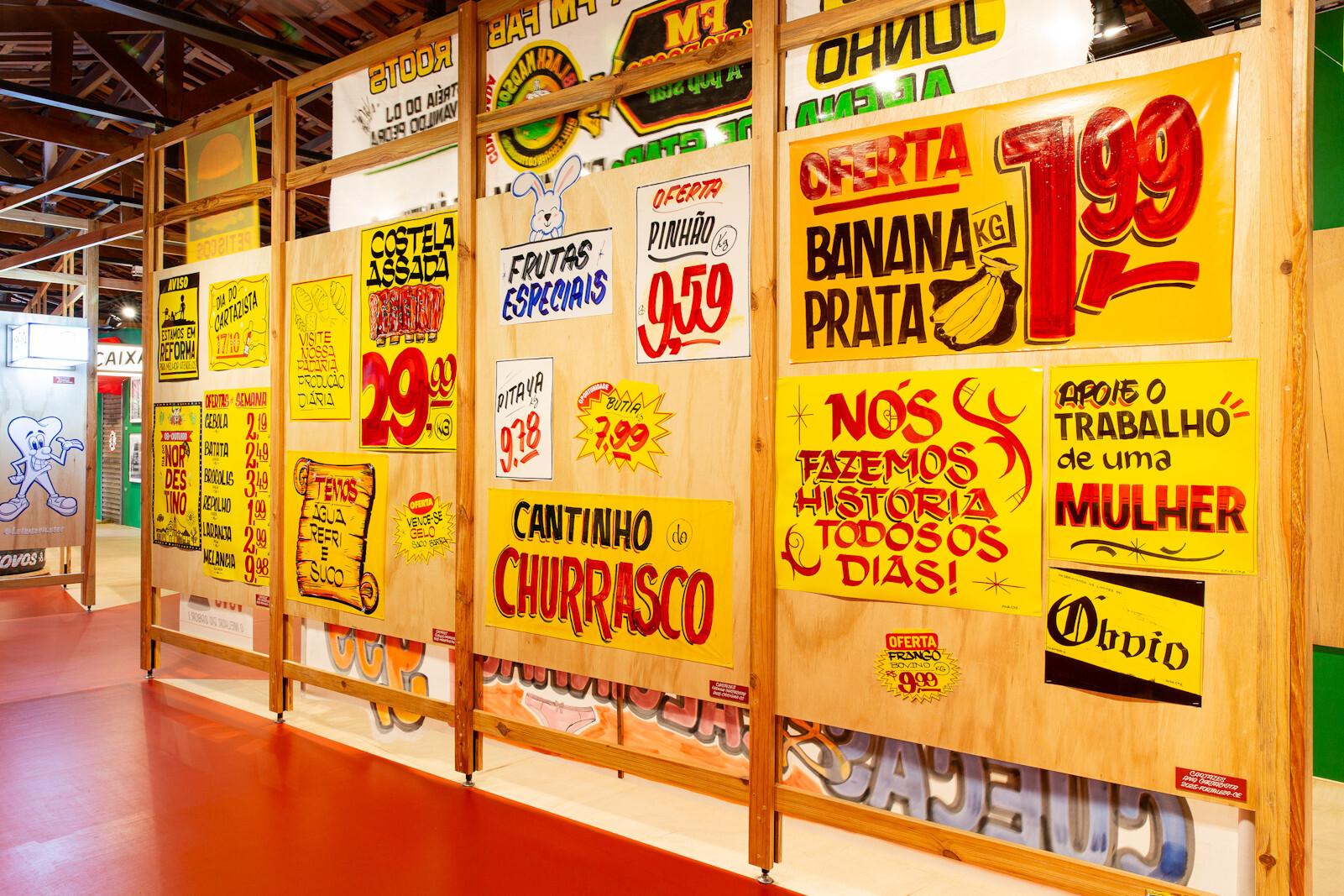 Hand-painted grocery store signs mounted to a series of display boards in a gallery setting. They are predominantly lettered in red on yellow backgrounds with a casual style coming from their ephemeral nature.