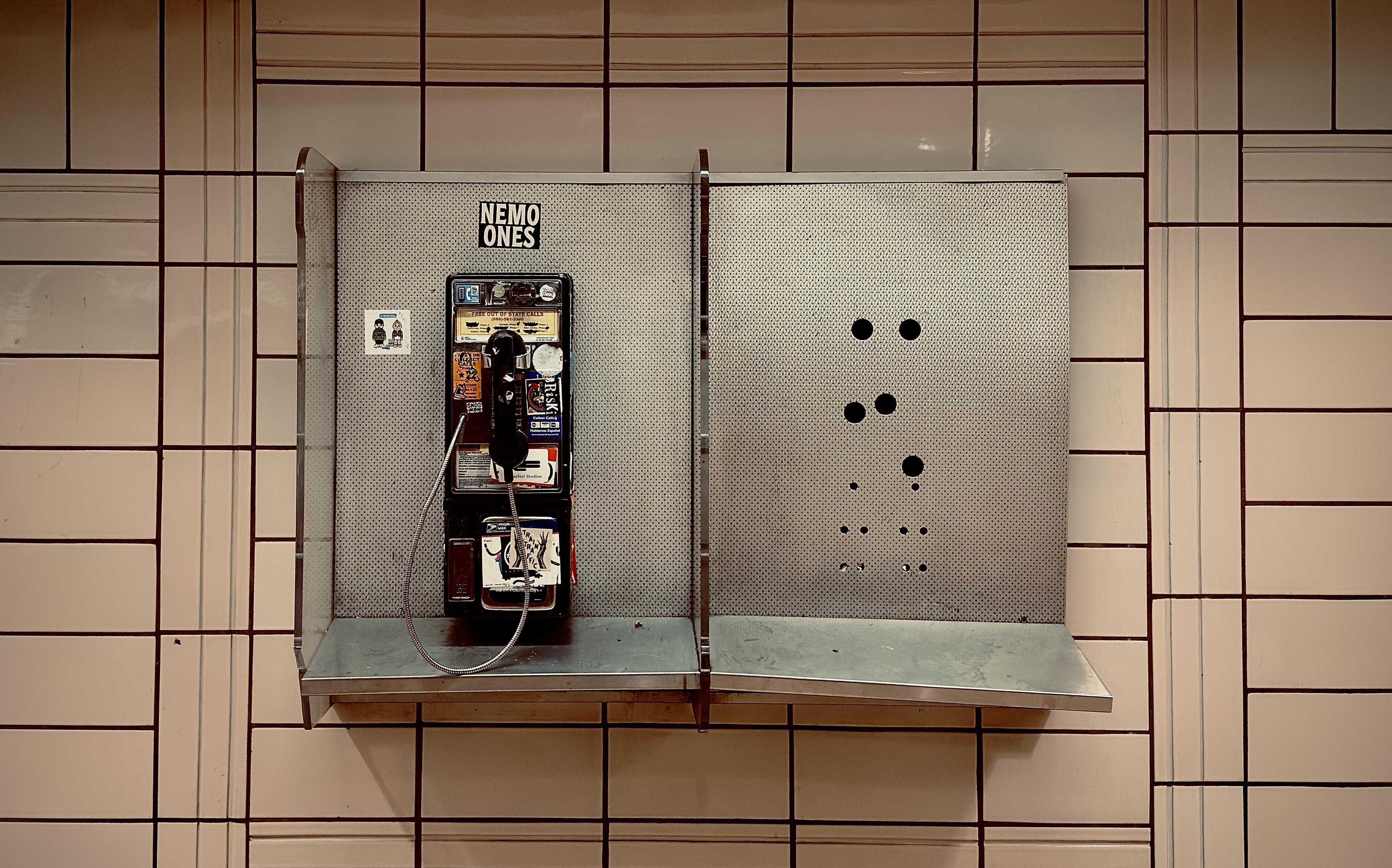 A color, medium-angle photograph in landscape orientation that captures a weathered, vintage pay phone mounted on a gridded tile wall within a pedestrian concourse beneath Rockefeller Center. The phone is housed in the left half of a dual-compartment, perforated metal booth with a narrow silver shelf running along the bottom. The black phone unit is heavily adorned with colorful stickers and graffiti tags, including a prominent "NEMO ONES" sticker centered directly above it. A metal-coiled handset cord loops down loosely, while the right compartment stands empty, revealing only a pattern of circular drill holes where a second phone was once installed. The backdrop consists of cream-colored rectangular tiles arranged in a distinct pattern of vertical and horizontal dark grout lines, characteristic of mid-century transit architecture. The lighting is warm and slightly dim, casting soft shadows beneath the metal shelf. The overall composition is symmetrical yet highlights a sense of urban decay and technological obsolescence, as the lonely, surviving phone sits next to the ghost of its former partner against the sterile, rhythmic pattern of the tiled wall.