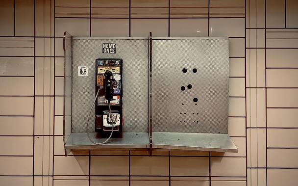A color, medium-angle photograph in landscape orientation that captures a weathered, vintage pay phone mounted on a gridded tile wall within a pedestrian concourse beneath Rockefeller Center. The phone is housed in the left half of a dual-compartment, perforated metal booth with a narrow silver shelf running along the bottom. The black phone unit is heavily adorned with colorful stickers and graffiti tags, including a prominent "NEMO ONES" sticker centered directly above it. A metal-coiled handset cord loops down loosely, while the right compartment stands empty, revealing only a pattern of circular drill holes where a second phone was once installed. The backdrop consists of cream-colored rectangular tiles arranged in a distinct pattern of vertical and horizontal dark grout lines, characteristic of mid-century transit architecture. The lighting is warm and slightly dim, casting soft shadows beneath the metal shelf. The overall composition is symmetrical yet highlights a sense of urban decay and technological obsolescence, as the lonely, surviving phone sits next to the ghost of its former partner against the sterile, rhythmic pattern of the tiled wall.