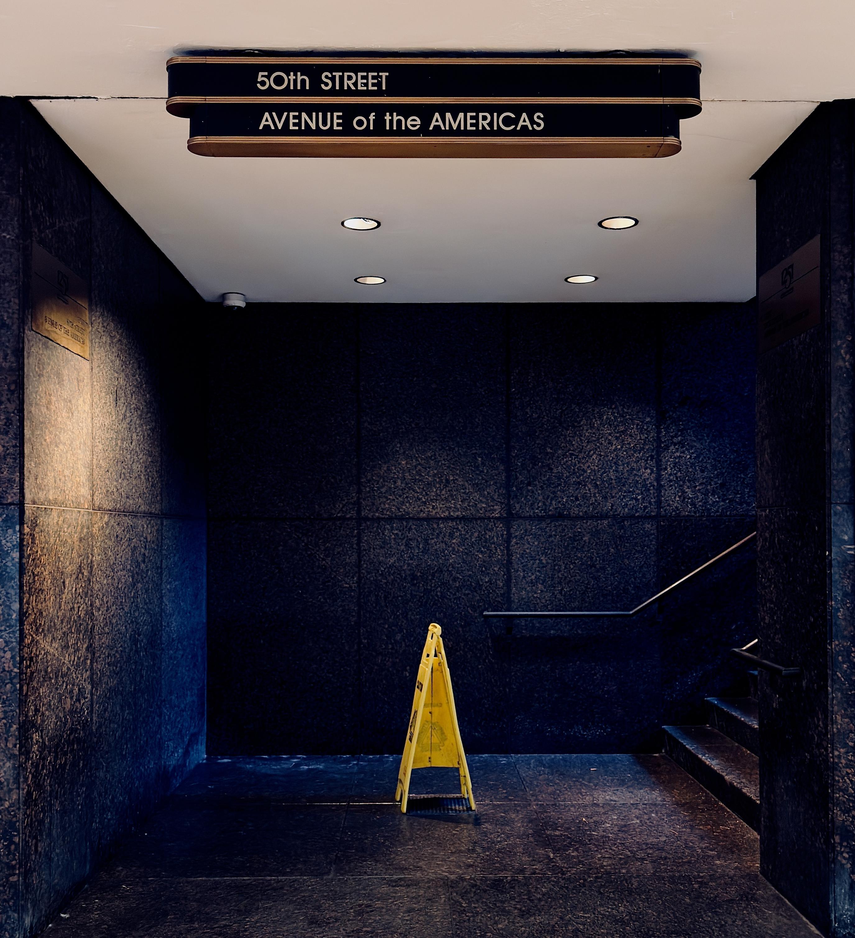 A color, medium-angle photograph in square aspect ratio that captures a dimly lit, moody interior corner of the underground pedestrian passageway at Rockefeller Center in Manhattan. The scene is dominated by dark, polished granite walls that meet at a sharp corner, with a staircase visible on the right side leading upward to the street level. In the center of the frame, a solitary yellow "Caution: Wet Floor" sign stands on the dark flooring, illuminated by four recessed circular lights in the white ceiling. The lighting creates a dramatic contrast, casting deep shadows in the corners while highlighting the texture of the stone and a silver metal handrail along the stairs. At the top of the frame, a prominent Art Deco-style sign hangs from the ceiling. The sign consists of two black horizontal tiers with gold-toned trim; the upper tier reads "50th STREET" and the lower tier reads "AVENUE of the AMERICAS" in clean, white capital letters. To the left, a brass plaque is mounted on the wall, partially caught in a warm glow from an unseen light source behind the sign.