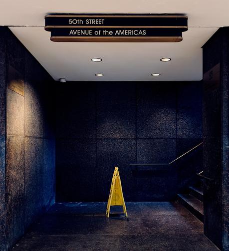 A color, medium-angle photograph in square aspect ratio that captures a dimly lit, moody interior corner of the underground pedestrian passageway at Rockefeller Center in Manhattan. The scene is dominated by dark, polished granite walls that meet at a sharp corner, with a staircase visible on the right side leading upward to the street level. In the center of the frame, a solitary yellow "Caution: Wet Floor" sign stands on the dark flooring, illuminated by four recessed circular lights in the white ceiling. The lighting creates a dramatic contrast, casting deep shadows in the corners while highlighting the texture of the stone and a silver metal handrail along the stairs. At the top of the frame, a prominent Art Deco-style sign hangs from the ceiling. The sign consists of two black horizontal tiers with gold-toned trim; the upper tier reads "50th STREET" and the lower tier reads "AVENUE of the AMERICAS" in clean, white capital letters. To the left, a brass plaque is mounted on the wall, partially caught in a warm glow from an unseen light source behind the sign.