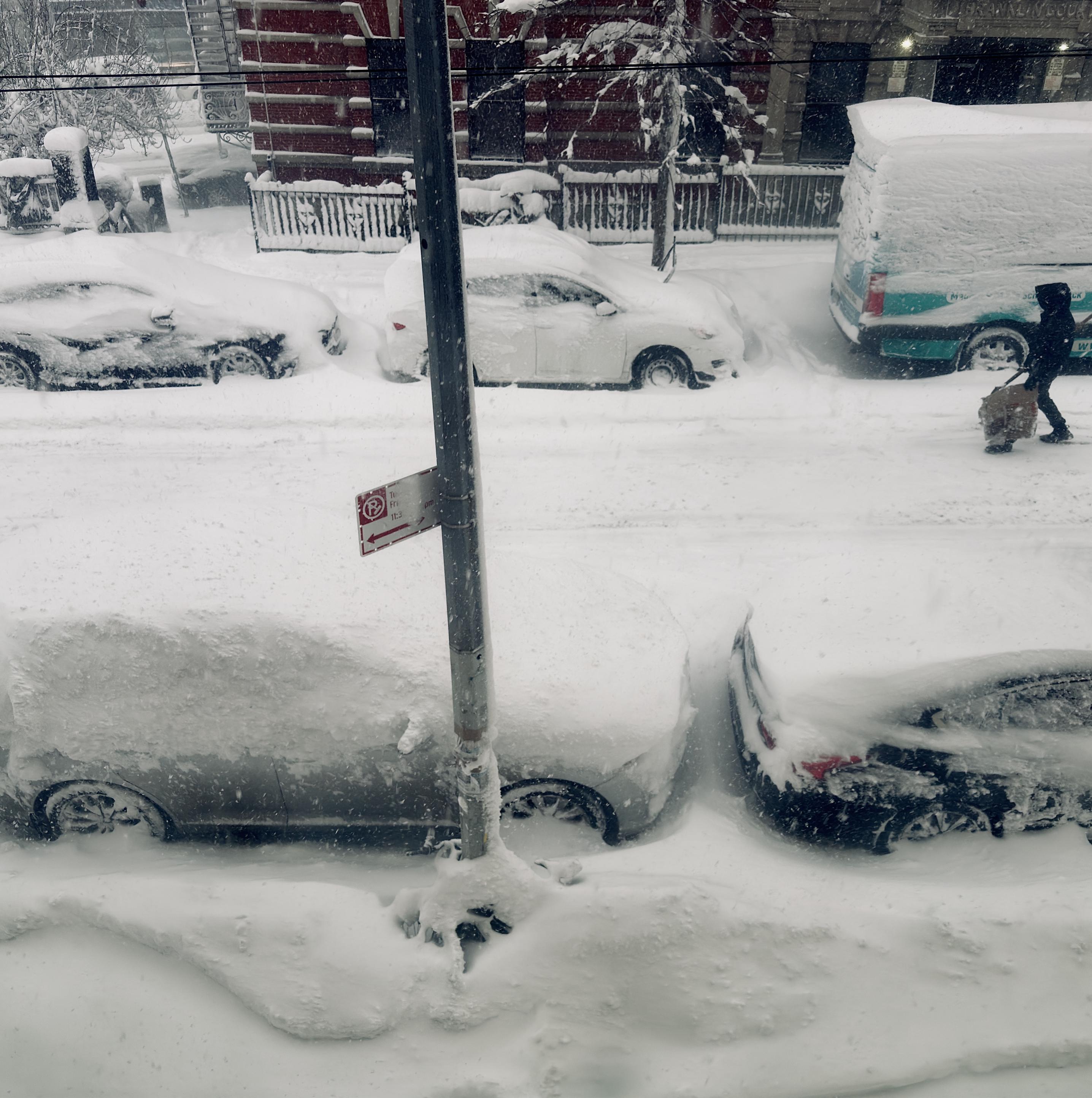 A color, medium-angle photograph taken from a high-angle that captures a residential street in Manhattan during a heavy winter storm. Several parked cars along both sides of the street are covered by deep, wind-swept snowdrifts, with their roofs, windshields, and hoods buried. A person dressed in dark winter gear is trekking their way down the middle of the street with a shovel, walking past a light blue and white delivery van. The red brick facade of a building and a black metal fence are partially visible behind the row of snow-covered vehicles on the far side of the street. In the center foreground, a grey metal utility pole stands tall, featuring a small red and white "No Parking" sign with an arrow pointing left. Chained to the base of this pole is a bicycle nearly entirely submerged in a massive mound of snow; the only visible part of the bike is the tip of a single handlebar poking out from the drift. The ground is a continuous, textured blanket of white, showing the sheer volume of the snowfall as it mounds up against the tires of the cars and the base of the buildings. Strong winds blow heavy falling snow through the overcast morning sky.