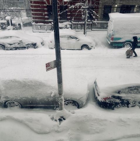 A color, medium-angle photograph taken from a high-angle that captures a residential street in Manhattan during a heavy winter storm. Several parked cars along both sides of the street are covered by deep, wind-swept snowdrifts, with their roofs, windshields, and hoods buried. A person dressed in dark winter gear is trekking their way down the middle of the street with a shovel, walking past a light blue and white delivery van. The red brick facade of a building and a black metal fence are partially visible behind the row of snow-covered vehicles on the far side of the street. In the center foreground, a grey metal utility pole stands tall, featuring a small red and white "No Parking" sign with an arrow pointing left. Chained to the base of this pole is a bicycle nearly entirely submerged in a massive mound of snow; the only visible part of the bike is the tip of a single handlebar poking out from the drift. The ground is a continuous, textured blanket of white, showing the sheer volume of the snowfall as it mounds up against the tires of the cars and the base of the buildings. Strong winds blow heavy falling snow through the overcast morning sky.