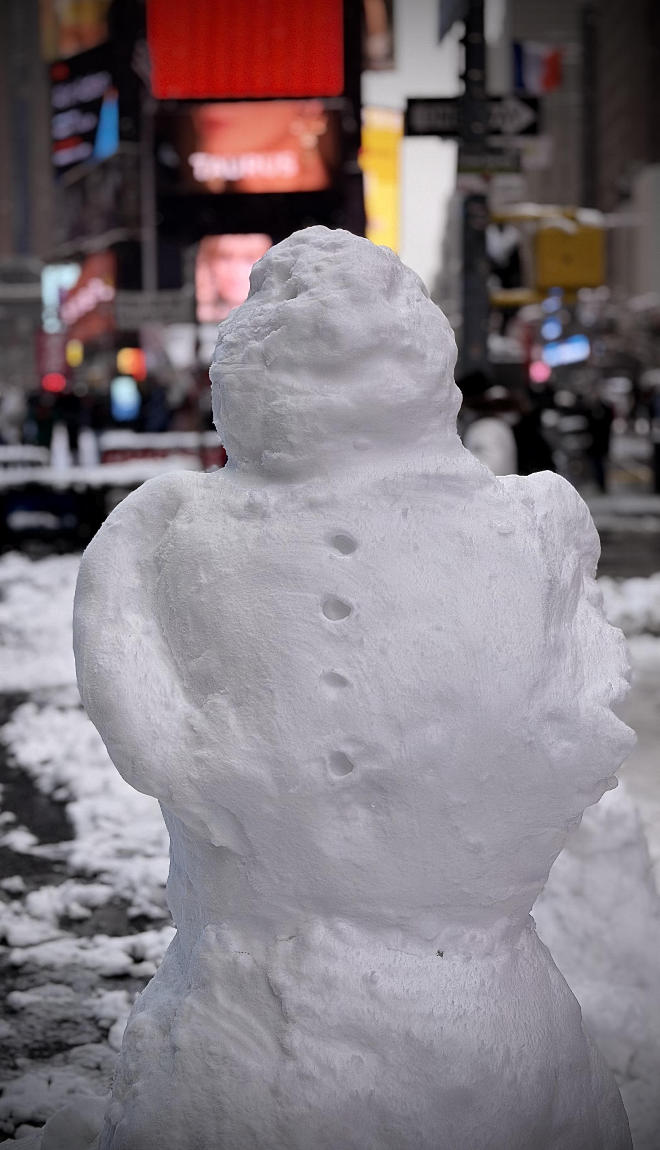 A color, medium-angle photograph in portrait orientation that captures a closeup of a hand-sculpted snowman standing in a snowy plaza, with the vibrant, blurred lights of Times Square in the background. The snowman is built from densely packed, slightly textured white snow and consists of three main sections. Its head is a rounded, somewhat lumpy orb, while its torso features four distinct indentations running vertically down the center to represent buttons. Small, arm-like mounds of snow are molded into the sides of the middle section, giving it a stout appearance. The background is intentionally out of focus, creating a bokeh effect that emphasizes the snowman as the central subject. Despite the blur, the iconic elements of New York City are visible, including bright red and yellow digital billboards, a street sign, and the faint silhouettes of pedestrians and city traffic. The ground is covered in a mixture of slush and fresh white snow. An overcast afternoon sky is evenly illuminating the scene.