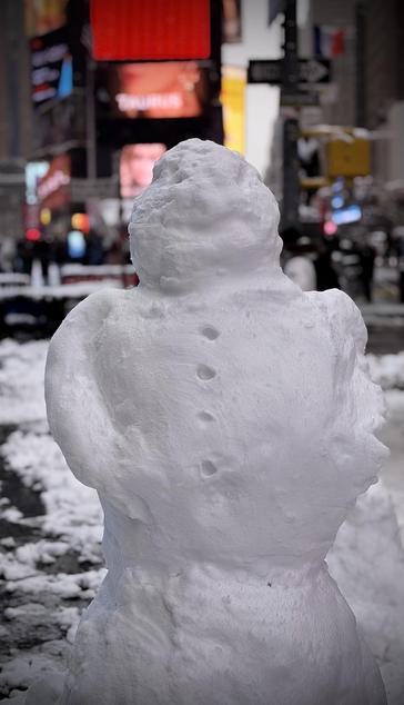 A color, medium-angle photograph in portrait orientation that captures a closeup of a hand-sculpted snowman standing in a snowy plaza, with the vibrant, blurred lights of Times Square in the background. The snowman is built from densely packed, slightly textured white snow and consists of three main sections. Its head is a rounded, somewhat lumpy orb, while its torso features four distinct indentations running vertically down the center to represent buttons. Small, arm-like mounds of snow are molded into the sides of the middle section, giving it a stout appearance. The background is intentionally out of focus, creating a bokeh effect that emphasizes the snowman as the central subject. Despite the blur, the iconic elements of New York City are visible, including bright red and yellow digital billboards, a street sign, and the faint silhouettes of pedestrians and city traffic. The ground is covered in a mixture of slush and fresh white snow. An overcast afternoon sky is evenly illuminating the scene.