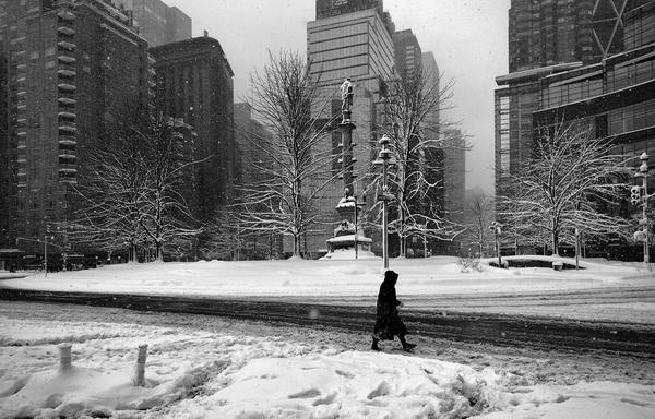 A noir, wide-angle photograph in landscape orientation that captures Columbus Circle in Manhattan during a heavy snowstorm. In the center of the frame, the iconic Christopher Columbus Monument stands atop its tall pedestal, flanked by skeletal, snow-covered trees that reach toward an overcast afternoon sky. A thick blanket of fresh snow covers the central plaza and the foreground, while a dark, slushy road curves through the middle ground, showing the tracks of passing vehicles exposing the asphalt. The surrounding skyscrapers, including the modern glass facade of the Deutsche Bank Center to the right, are partially obscured by the heavy falling snow. In the lower-right foreground, a lone pedestrian dressed in a long, dark coat and hood walks across the slushy street, their silhouette providing a sharp contrast against the white landscape. The composition is filled with the texture of falling snowflakes, which appear as soft white specks against the darker buildings and trees. The overall mood is quiet and atmospheric, emphasizing the stillness and cold of a city momentarily slowed by the elements.