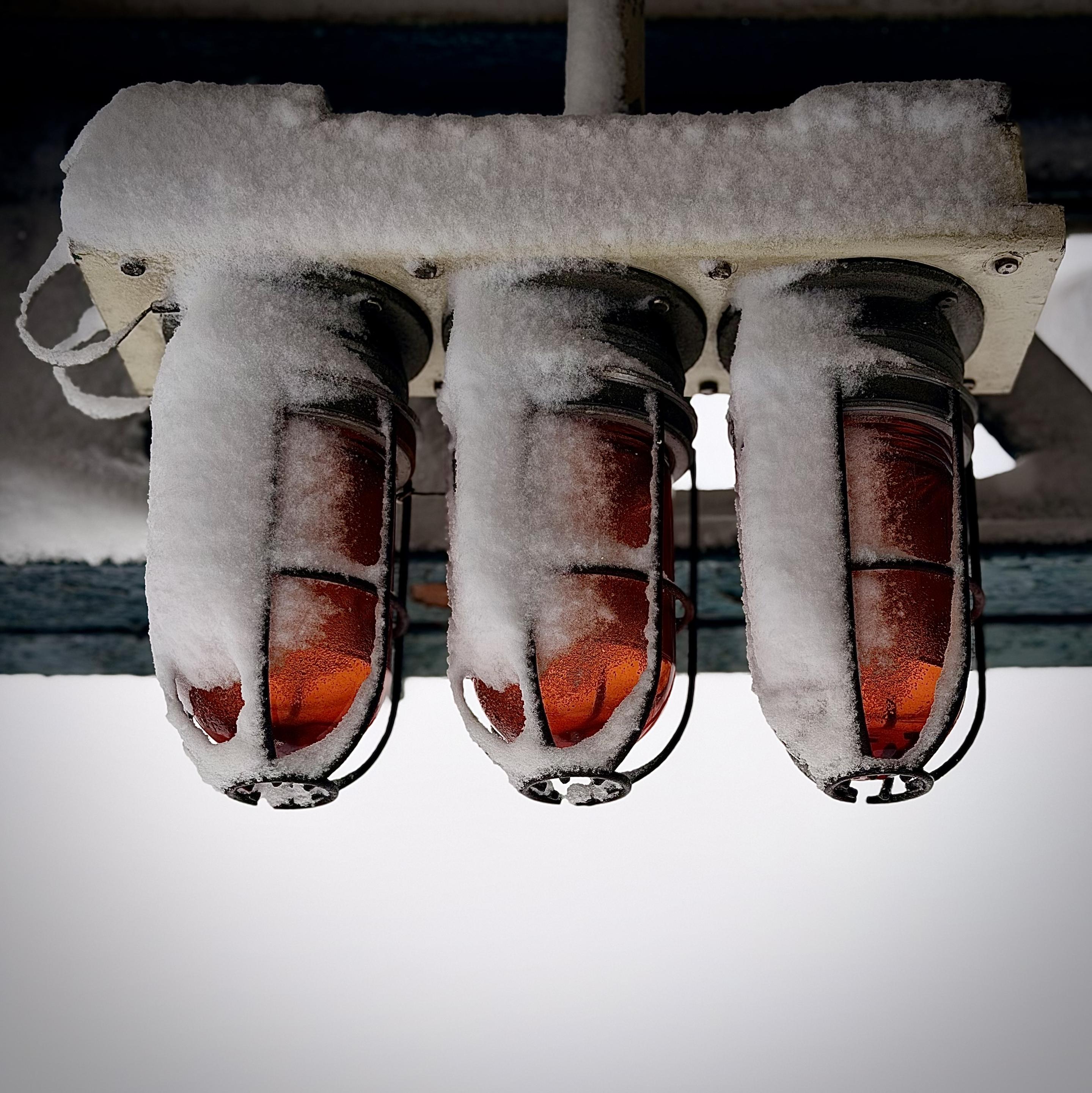 A color, low-angle photograph in square aspect ratio that captures a set of three industrial-style signal lights at Prospect Avenue Station in the Bronx after a snowstorm. The three lights are encased in rounded, amber-colored glass globes, each protected by a black metal wire cage. A thick, textured layer of white snow blankets the flat rectangular base of the fixture and heavily clings to the top and left sides of each light, creating frozen, hood-like shapes. The amber glass glows with a warm, deep orange hue, which contrasts sharply against the stark white of the snow and the overcast, pale background. The perspective is looking upward toward the ceiling of the station platform, with a dark, weathered beam visible just above the fixture. The snow appears heavy and damp, partially obscuring the metal cages and the top halves of the glowing globes. Below the lights, the scene fades into a bright, hazy white void of the afternoon winter sky. The overall mood is cold and industrial.