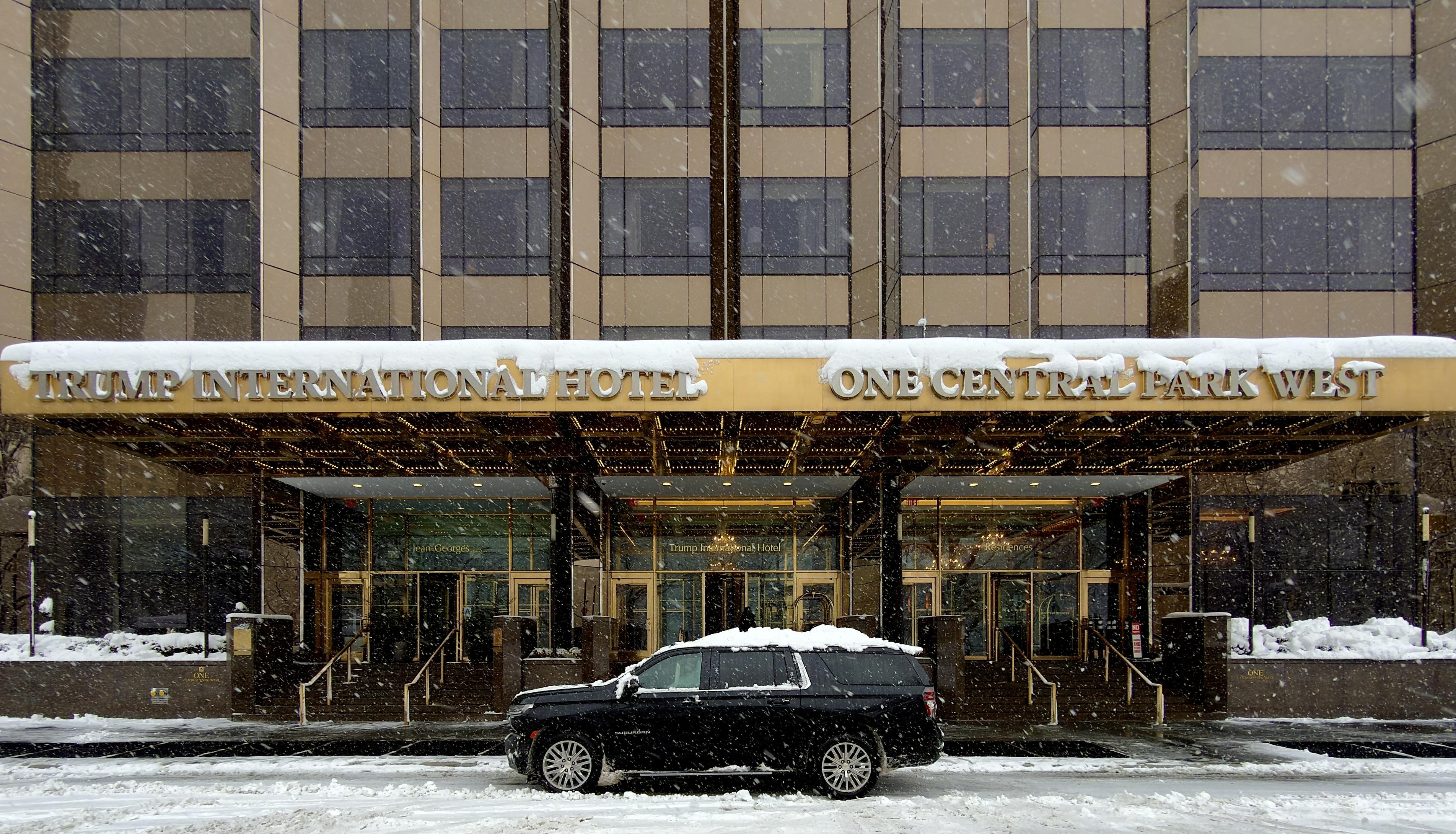 A color, wide-angle photograph in landscape orientation that captures an eye-level shot of the exterior entrance of the Trump International Hotel at One Central Park West during a heavy winter storm. The building’s facade features reflective dark glass panels framed by a metallic, gold-bronze colored grid. A prominent, wide gold awning stretches across the front of the hotel, heavily capped with a thick layer of fresh white snow. In bold, dark lettering, the awning reads "TRUMP INTERNATIONAL HOTEL" on the left and "ONE CENTRAL PARK WEST" on the right. The lettering is also capped with accumulating snow. Below the overhang, the revolving glass doors and warm interior lights of the lobby are visible, contrasted against the cold, gray atmosphere of the blizzard. In the foreground, a black SUV is parked parallel to the building on the street blanketed in slush and deep snow. The vehicle's roof and hood are partially covered in white, blending into the wintry scene. Thick snowflakes fill the air, creating a hazy, textured veil across the entire frame. The sidewalks and the low stone walls bordering the entrance are buried under several inches of snow, and the overall color palette is dominated by muted grays, whites, and the dull glint of the gold architectural accents underneath the overcast morning sky.