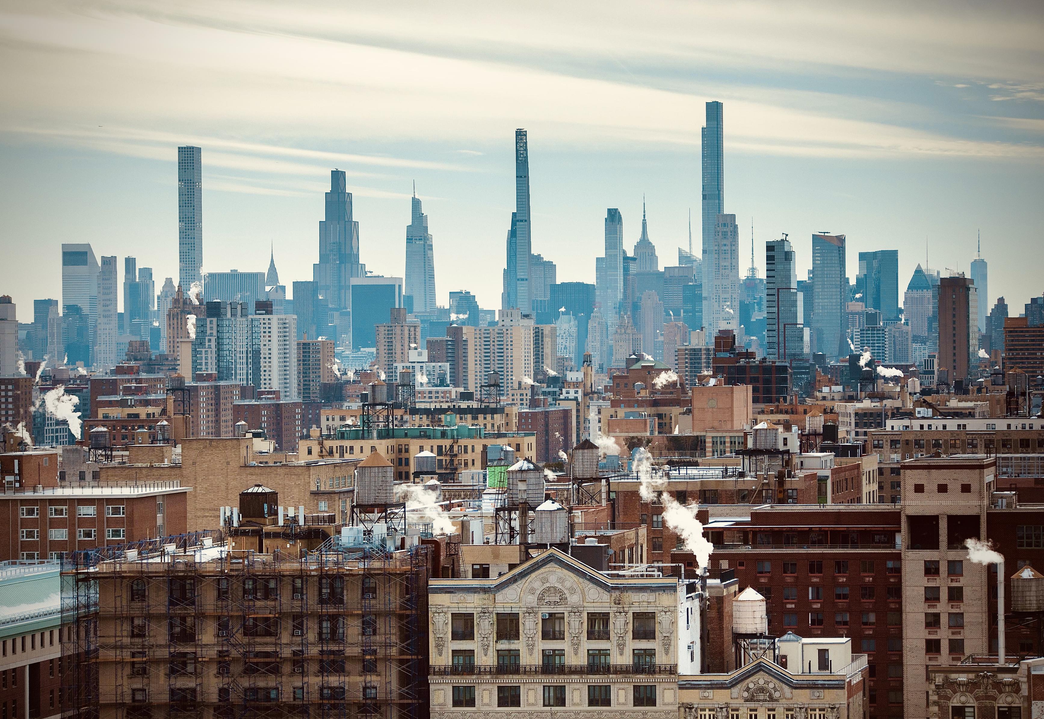 A color, wide-angle photograph taken from a high vantage point captures a south-facing view of the Manhattan skyline landscape on a crisp, partly overcast day. In the foreground and midground, the architecture of the Upper West Side is defined by a sea of tan, red-brick, and beige pre-war apartment buildings. These structures are topped with iconic wooden water towers and numerous chimneys emitting white plumes of steam that drift into the pale sky. The perspective creates a layered effect, where the older, shorter residential blocks transition into the more modern, glass-clad mid-rise buildings of the Midtown district. In the background, the horizon is dominated by the skyscrapers of midtown Manhattan. Notable landmarks include the ultra-slender silhouettes of 432 Park Avenue, 111 West 57th Street (Steinway Tower), and the Central Park Tower. To the right, the Art Deco spire of the Empire State Building peeks out from behind newer glassy towers, while the distant, pointed needle of One World Trade Center is visible on the far right horizon. The color palette is a muted mix of earthy brick tones and cool blue-gray glass, unified by a soft, diffused sunlight that casts gentle shadows across the city grid.