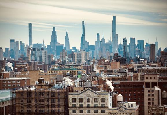 A color, wide-angle photograph taken from a high vantage point captures a south-facing view of the Manhattan skyline landscape on a crisp, partly overcast day. In the foreground and midground, the architecture of the Upper West Side is defined by a sea of tan, red-brick, and beige pre-war apartment buildings. These structures are topped with iconic wooden water towers and numerous chimneys emitting white plumes of steam that drift into the pale sky. The perspective creates a layered effect, where the older, shorter residential blocks transition into the more modern, glass-clad mid-rise buildings of the Midtown district. In the background, the horizon is dominated by the skyscrapers of midtown Manhattan. Notable landmarks include the ultra-slender silhouettes of 432 Park Avenue, 111 West 57th Street (Steinway Tower), and the Central Park Tower. To the right, the Art Deco spire of the Empire State Building peeks out from behind newer glassy towers, while the distant, pointed needle of One World Trade Center is visible on the far right horizon. The color palette is a muted mix of earthy brick tones and cool blue-gray glass, unified by a soft, diffused sunlight that casts gentle shadows across the city grid.