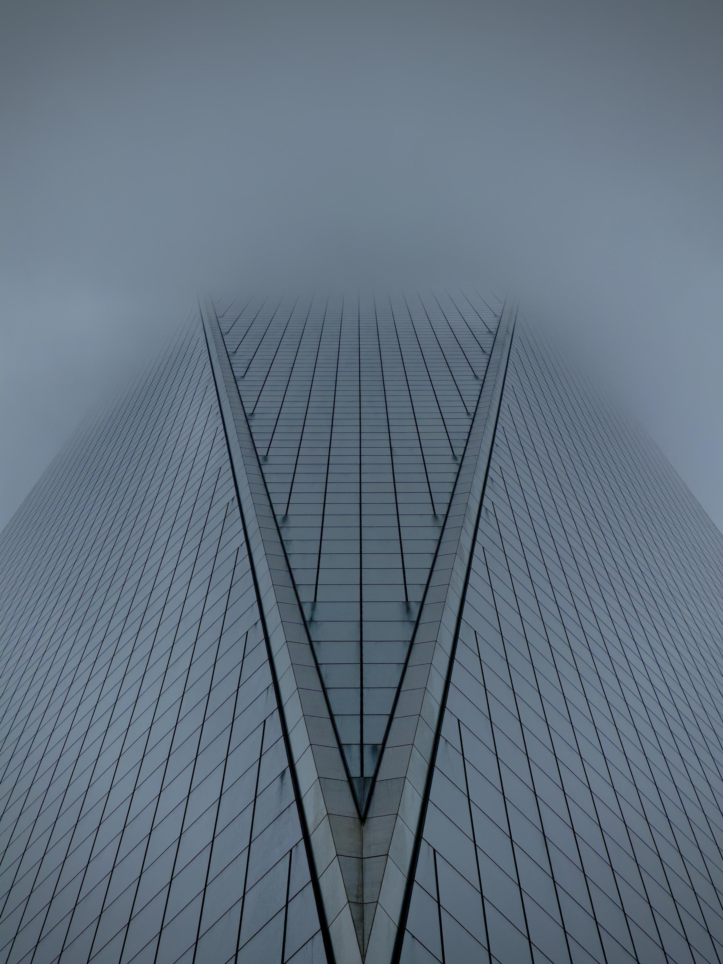 A color, low-angle, symmetrical photograph in portrait orientation that captures the towering facade of One World Trade Center as it ascends directly into a thick, heavy shroud of grey fog on a rainy morning in lower Manhattan. The perspective emphasizes the building's geometric precision, focusing on the prominent V-shaped indentation of its glass curtain wall. This architectural feature creates a sharp, converging point at the bottom center of the frame, with the reflective glass panels forming a rhythmic grid of vertical and diagonal lines that lead the eye upward. The color palette is monochromatic and somber, dominated by cool slate blues and muted silvers that mirror the gloomy, overcast sky. The upper portion of the skyscraper gradually dissolves into a dense mist, rendering the top of the building invisible and creating an ethereal, infinite quality. The lighting is soft and diffused, typical of a rainy day, which eliminates harsh shadows and highlights the sleek, metallic texture of the window frames. Fine water droplets and a misty haze add a soft-focus effect to the glass, reinforcing the damp, atmospheric mood of the scene. The overall composition is minimalist and imposing, stripping away the surrounding city to focus entirely on the building’s interaction with the elements.
