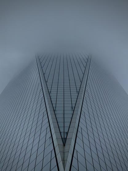 A color, low-angle, symmetrical photograph in portrait orientation that captures the towering facade of One World Trade Center as it ascends directly into a thick, heavy shroud of grey fog on a rainy morning in lower Manhattan. The perspective emphasizes the building's geometric precision, focusing on the prominent V-shaped indentation of its glass curtain wall. This architectural feature creates a sharp, converging point at the bottom center of the frame, with the reflective glass panels forming a rhythmic grid of vertical and diagonal lines that lead the eye upward. The color palette is monochromatic and somber, dominated by cool slate blues and muted silvers that mirror the gloomy, overcast sky. The upper portion of the skyscraper gradually dissolves into a dense mist, rendering the top of the building invisible and creating an ethereal, infinite quality. The lighting is soft and diffused, typical of a rainy day, which eliminates harsh shadows and highlights the sleek, metallic texture of the window frames. Fine water droplets and a misty haze add a soft-focus effect to the glass, reinforcing the damp, atmospheric mood of the scene. The overall composition is minimalist and imposing, stripping away the surrounding city to focus entirely on the building’s interaction with the elements.