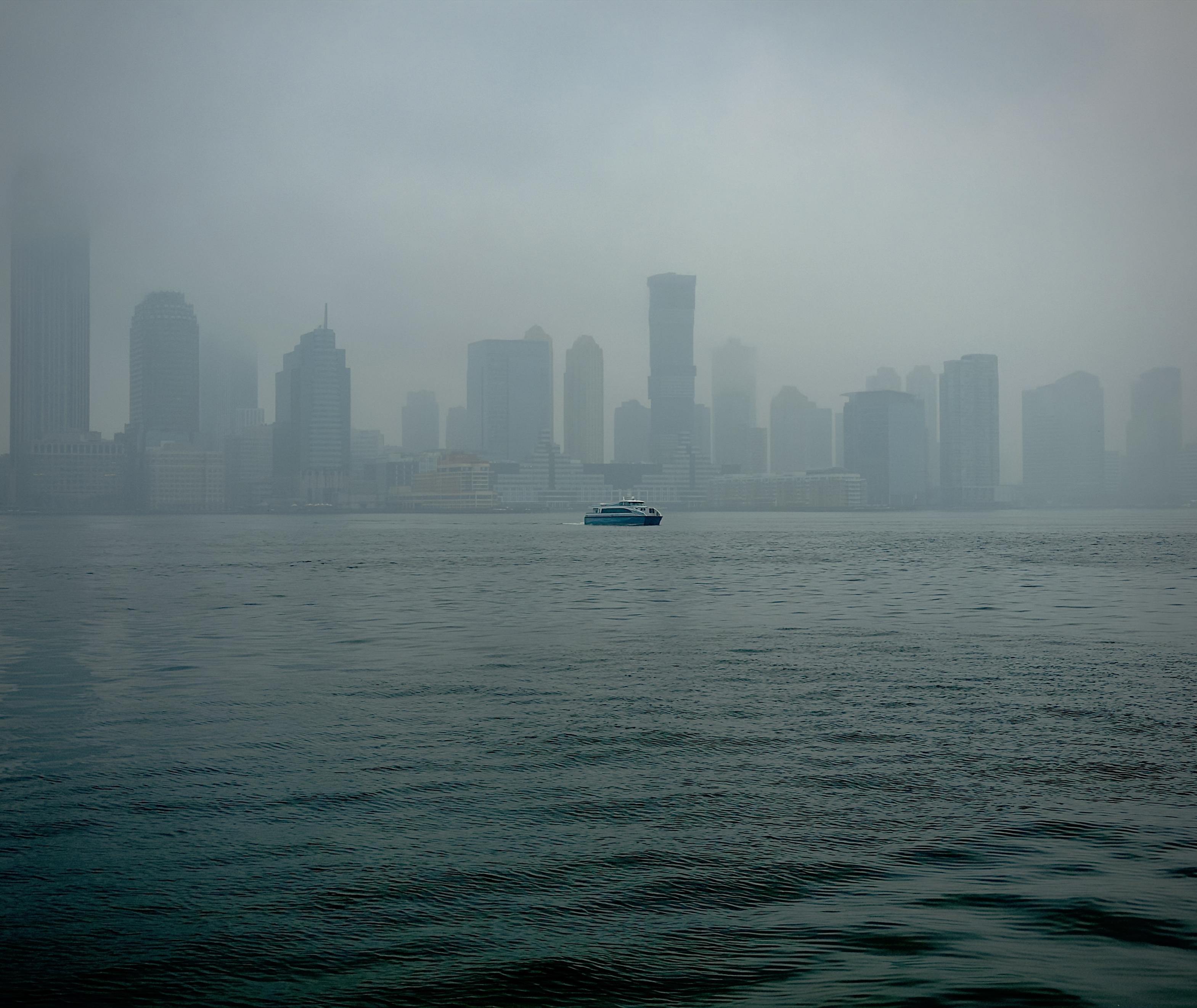 A color, wide-angle photograph that captures a view of the Jersey City skyline as seen from across the Hudson River in lower Manhattan on a heavily overcast, foggy day. The water in the foreground is a deep, moody teal with subtle, rhythmic ripples moving across the surface. Centered in the middle distance, a small blue and white ferry cuts through the water, providing a sharp point of contrast against the muted surroundings. The thick fog hangs low over the river, partially obscuring the base of the buildings and softening the horizon line where the water meets the city. The skyline itself is composed of several high-rise office and residential towers, rendered in varying shades of grey and pale blue. Due to the dense mist, the buildings toward the edges of the frame appear as ghostly silhouettes, while the more central structures show a bit more architectural detail and less muted colors. The sky is a seamless, flat expanse of light grey fog that blends into the tops of the tallest skyscrapers, creating a quiet, atmospheric urban landscape.