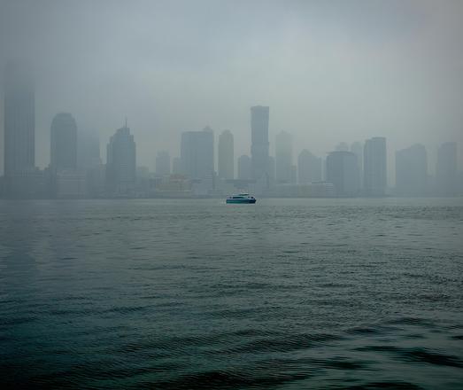 A color, wide-angle photograph that captures a view of the Jersey City skyline as seen from across the Hudson River in lower Manhattan on a heavily overcast, foggy day. The water in the foreground is a deep, moody teal with subtle, rhythmic ripples moving across the surface. Centered in the middle distance, a small blue and white ferry cuts through the water, providing a sharp point of contrast against the muted surroundings. The thick fog hangs low over the river, partially obscuring the base of the buildings and softening the horizon line where the water meets the city. The skyline itself is composed of several high-rise office and residential towers, rendered in varying shades of grey and pale blue. Due to the dense mist, the buildings toward the edges of the frame appear as ghostly silhouettes, while the more central structures show a bit more architectural detail and less muted colors. The sky is a seamless, flat expanse of light grey fog that blends into the tops of the tallest skyscrapers, creating a quiet, atmospheric urban landscape.