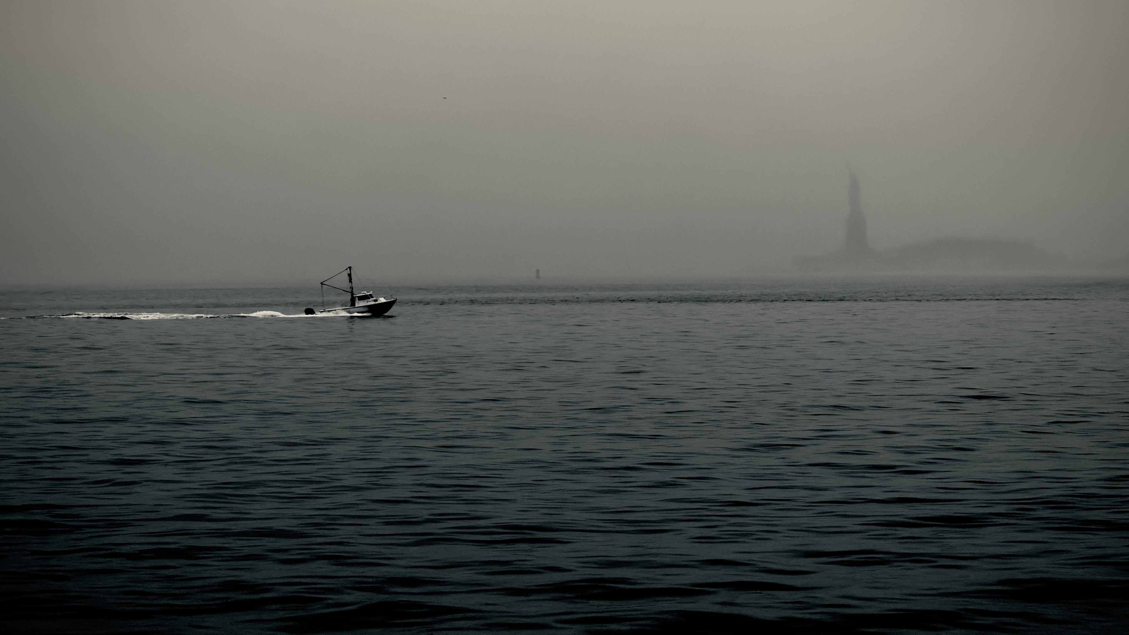 A noir, wide-angle photograph in landscape orientation that captures a moody view of Upper New York Bay on a heavily overcast and foggy day. The water in the foreground is dark and choppy, with small, rhythmic waves rippling across the surface. To the left, a small white and dark-hulled tug boat moves across the mid-ground from the left side of the frame, leaving a distinct white wake in its path. The atmosphere is thick with mist, blurring the horizon line where the gray sea meets a pale, uniform sky. In the far distance to the right, the Statue of Liberty appears as a ghostly, dark silhouette. The heavy fog leaving only its iconic torch-bearing shape visible against the hazy backdrop of Liberty Island. The entire scene is rendered in a desaturated palette, emphasizing the sense of solitude and quiet.