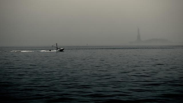 A noir, wide-angle photograph in landscape orientation that captures a moody view of Upper New York Bay on a heavily overcast and foggy day. The water in the foreground is dark and choppy, with small, rhythmic waves rippling across the surface. To the left, a small white and dark-hulled tug boat moves across the mid-ground from the left side of the frame, leaving a distinct white wake in its path. The atmosphere is thick with mist, blurring the horizon line where the gray sea meets a pale, uniform sky. In the far distance to the right, the Statue of Liberty appears as a ghostly, dark silhouette. The heavy fog leaving only its iconic torch-bearing shape visible against the hazy backdrop of Liberty Island. The entire scene is rendered in a desaturated palette, emphasizing the sense of solitude and quiet.