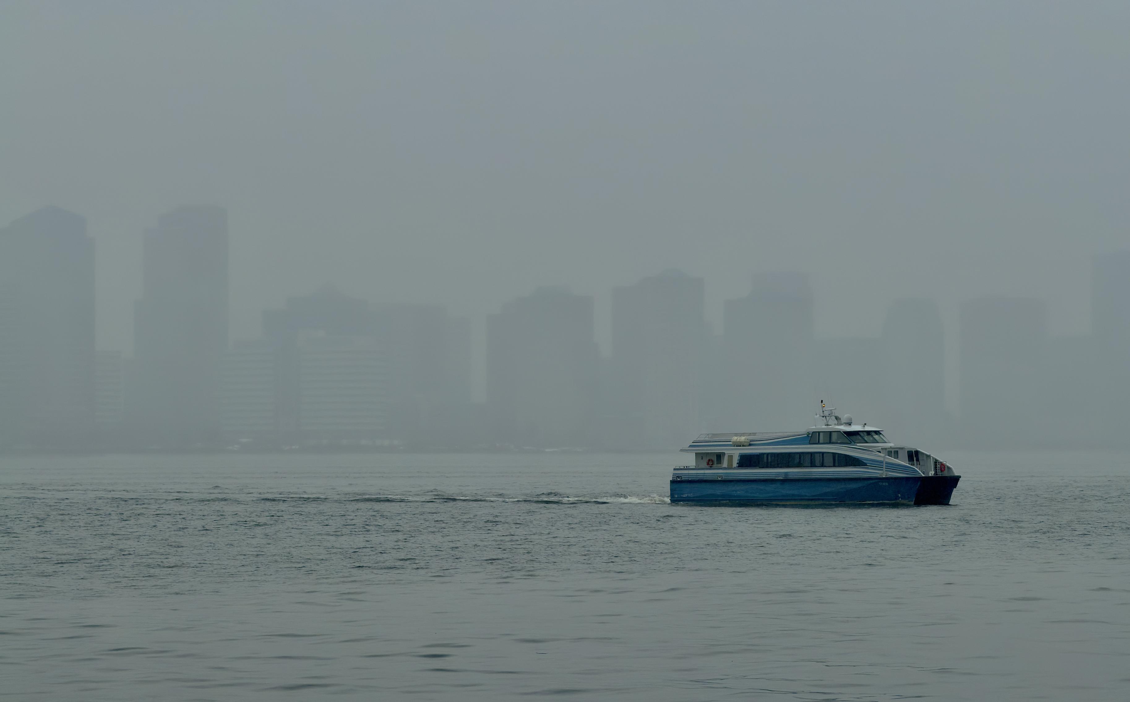 A color, wide-angle photograph in landscape orientation that captures the Hudson River on a densely foggy afternoon. A side-profile view of a blue and white catamaran ferry gliding across the gray, choppy waters in the lower-right quadrant is the sharpest element in the frame, creating a white wake behind it as it moves from right to left. The boat features a sleek, modern design with two visible decks and dark-tinted windows running along its length. The background consists of the Jersey City skyline, which is heavily obscured by a dense, monochromatic shroud of fog and haze. The skyscrapers appear as faint, ghostly silhouettes in various shades of muted gray, lacking any sharp architectural detail. The overcast sky and the water blend into a seamless, atmospheric gradient, emphasizing a gloomy, rainy, and tranquil mood through a limited color palette of cool blues and grays.