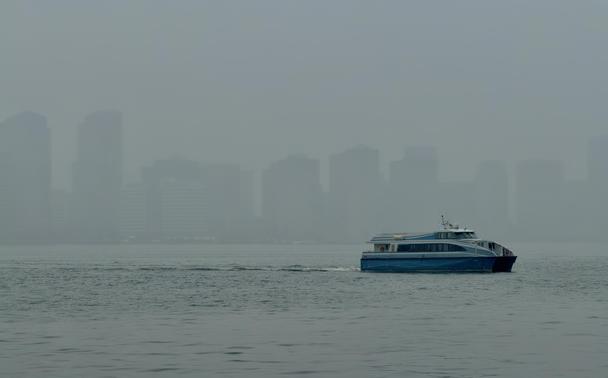 A color, wide-angle photograph in landscape orientation that captures the Hudson River on a densely foggy afternoon. A side-profile view of a blue and white catamaran ferry gliding across the gray, choppy waters in the lower-right quadrant is the sharpest element in the frame, creating a white wake behind it as it moves from right to left. The boat features a sleek, modern design with two visible decks and dark-tinted windows running along its length. The background consists of the Jersey City skyline, which is heavily obscured by a dense, monochromatic shroud of fog and haze. The skyscrapers appear as faint, ghostly silhouettes in various shades of muted gray, lacking any sharp architectural detail. The overcast sky and the water blend into a seamless, atmospheric gradient, emphasizing a gloomy, rainy, and tranquil mood through a limited color palette of cool blues and grays.
