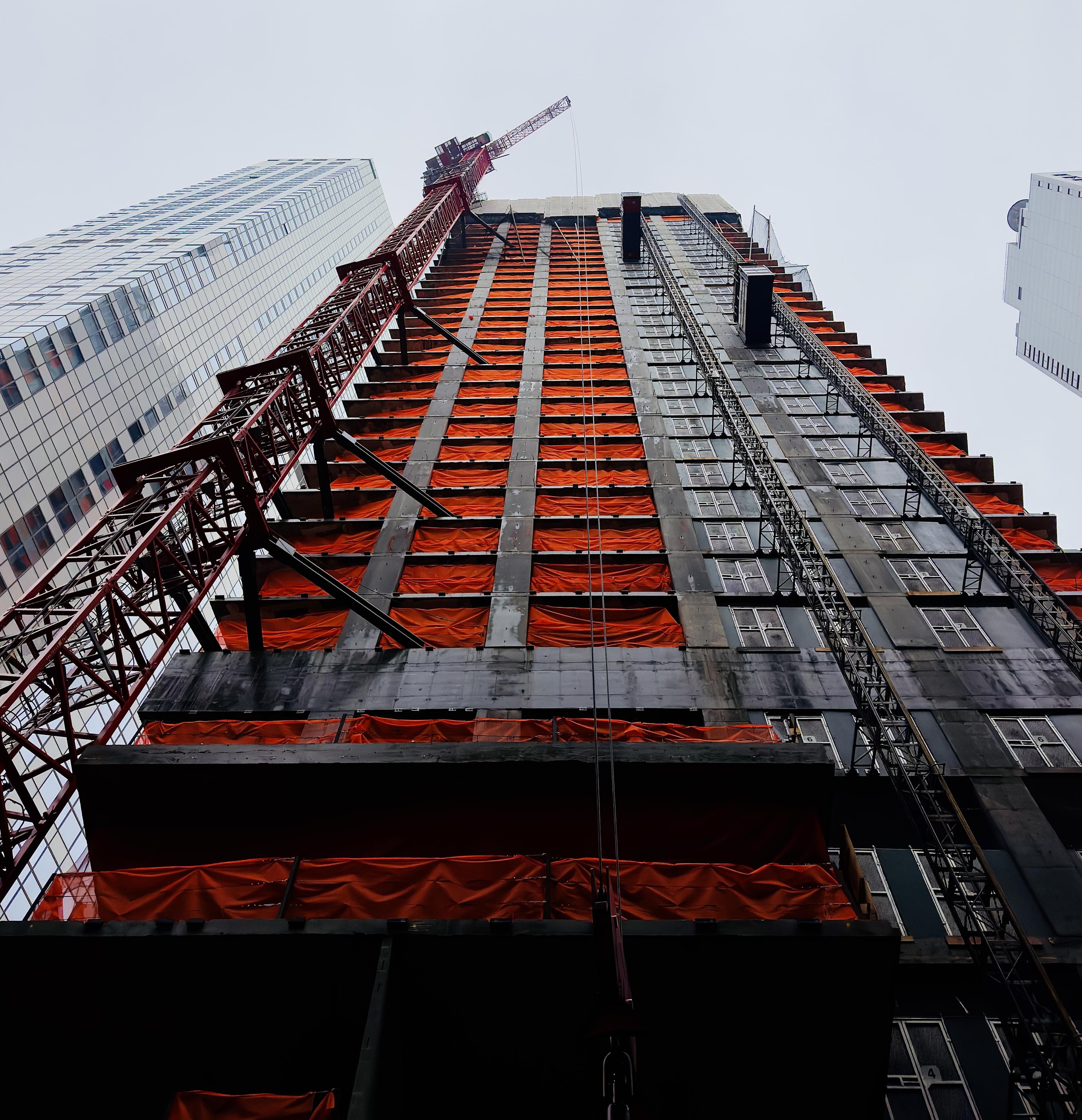 A color, low-angle photograph in portrait orientation that captures a skyscraper under construction in the Financial District of Manhattan. The building appears soaring upward against a pale, overcast winter sky. The building’s concrete skeletal frame is prominently draped in bright orange safety netting that covers nearly every floor, creating a rhythmic pattern of vibrant color against the dark, unfinished exterior. To the left, a massive red lattice crane is bolted to the side of the structure, its arm reaching toward the top, while a mechanical construction hoist runs vertically along the right side of the facade. The perspective emphasizes the sheer height and industrial scale of the project, framed by the finished glass and steel facades of neighboring skyscrapers on either side. In the foreground, the lower levels of the building are shrouded in deep shadow, contrasting with the light hitting the upper floors. The scene is a dense composition of geometric lines, featuring the grid of the window openings, the diagonal braces of the crane, and the metallic texture of the external elevators. The a scene is a stark contrast between the rich orange safety netting and the cool grays and blues that fill the frame.