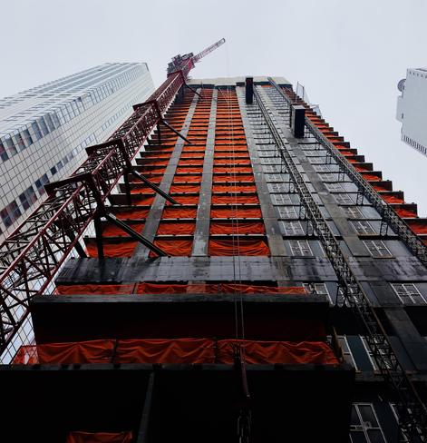 A color, low-angle photograph in portrait orientation that captures a skyscraper under construction in the Financial District of Manhattan. The building appears soaring upward against a pale, overcast winter sky. The building’s concrete skeletal frame is prominently draped in bright orange safety netting that covers nearly every floor, creating a rhythmic pattern of vibrant color against the dark, unfinished exterior. To the left, a massive red lattice crane is bolted to the side of the structure, its arm reaching toward the top, while a mechanical construction hoist runs vertically along the right side of the facade. The perspective emphasizes the sheer height and industrial scale of the project, framed by the finished glass and steel facades of neighboring skyscrapers on either side. In the foreground, the lower levels of the building are shrouded in deep shadow, contrasting with the light hitting the upper floors. The scene is a dense composition of geometric lines, featuring the grid of the window openings, the diagonal braces of the crane, and the metallic texture of the external elevators. The a scene is a stark contrast between the rich orange safety netting and the cool grays and blues that fill the frame.