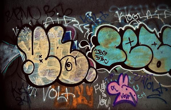 A color, medium-angle photograph in landscape orientation that captures a vibrant and textured wall of urban street art. The wall is covered in a dense layer of overlapping graffiti tags and large throw-up style pieces. On the left, a rounded, bubble-letter piece in pale yellow and cream features thick black outlines and subtle blue highlights, while to the right, a similar piece is filled with a weathered turquoise and seafoam green gradient. White and blue scrawled handstyles, including the recurring name "VOLT," and "KOOL AID" fill the negative space around the larger letters. In the lower right corner, a small, whimsical character resembling a pink rabbit head with a purple outline adds a pop of bright color against the dark, gritty background, illustrating the complex, layered history of the mural.
