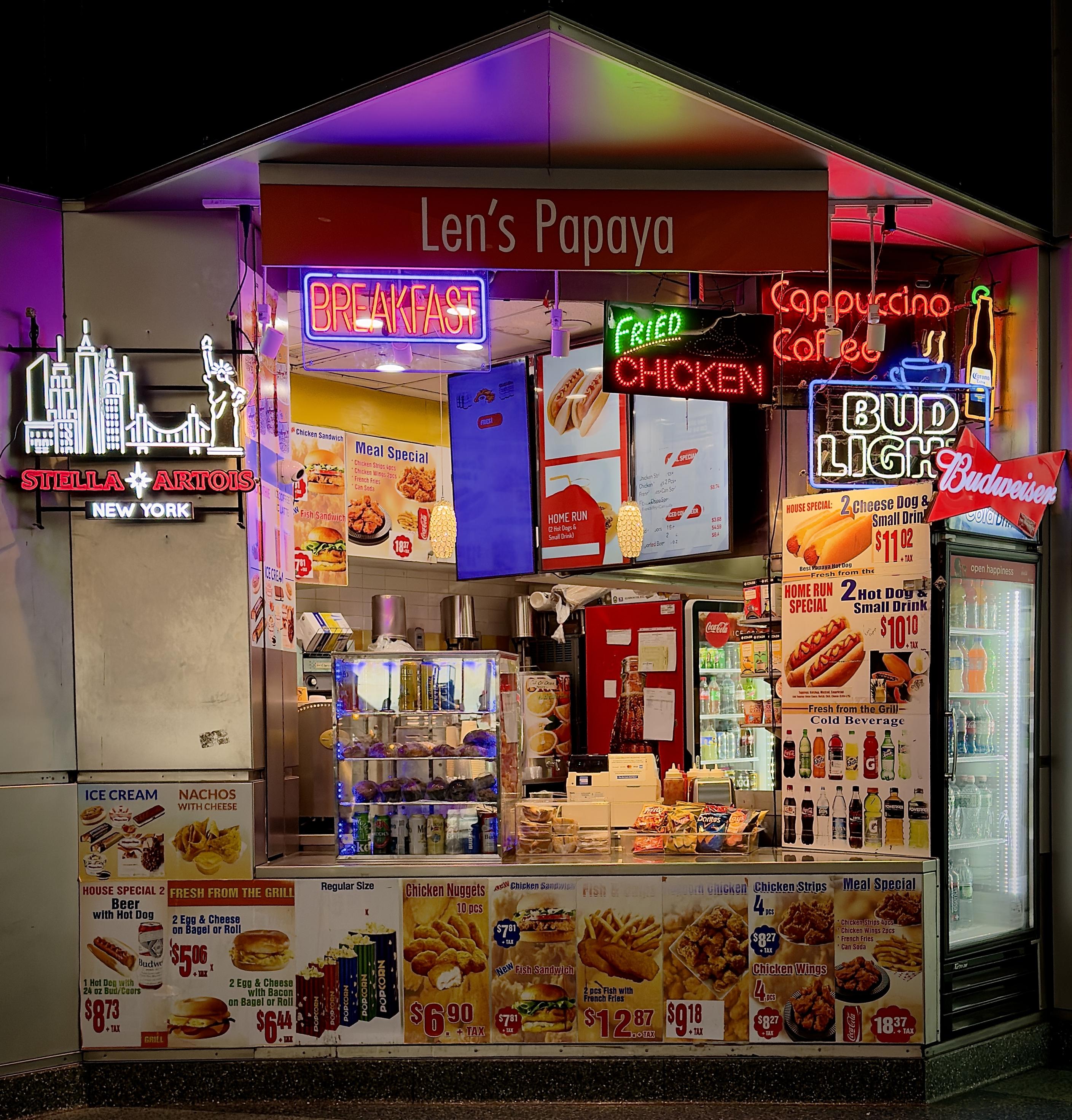 A color, medium-angle photograph in square aspect ratio that captures "Len’s Papaya” inside of Whitehall Terminal in lower Manhattan. The stall is a brightly lit food kiosk featuring a vibrant array of neon signs and colorful menu advertisements. To the left, a large neon sign depicts the New York City skyline with the Statue of Liberty and the Brooklyn Bridge above a "Stella Artois New York" logo. The kiosk's facade is densely packed with glowing signs for "BREAKFAST," "FRIED CHICKEN," "Cappuccino Coffee," and "BUD LIGHT,". A large "Budweiser" bowtie logo is perched on the corner of a glass-door beverage refrigerator filled with bottled sodas and water. Below the counter, a series of detailed menu posters display various food offerings, including hot dogs, chicken nuggets, fish and chips, and breakfast sandwiches. Notable specials listed include a "Home Run Special" with two hot dogs and a small drink, and a beer with a hot dog combo. The interior of the kiosk reveals a densely packed workspace with beverage dispensers, a clear pastry case filled with muffins, and shelves stocked with snacks like chips. The lighting is a mix of warm overhead bulbs and the colorful, multi-colored glow from the neon signs, reflecting off the metallic and glass surfaces of the stand. The largest sign atop the space says “Len’s Papaya” in white lettering on a red background.