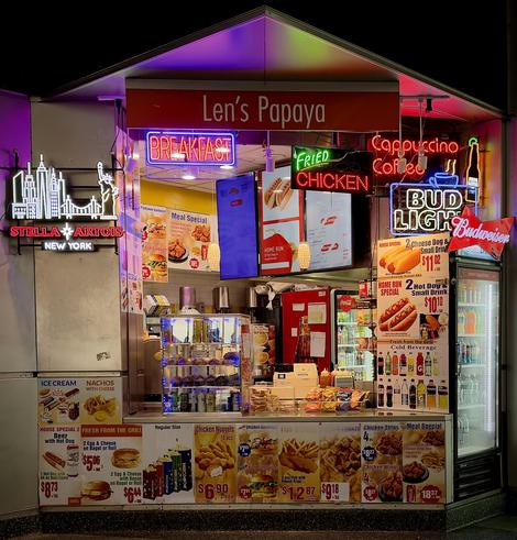 A color, medium-angle photograph in square aspect ratio that captures "Len’s Papaya” inside of Whitehall Terminal in lower Manhattan. The stall is a brightly lit food kiosk featuring a vibrant array of neon signs and colorful menu advertisements. To the left, a large neon sign depicts the New York City skyline with the Statue of Liberty and the Brooklyn Bridge above a "Stella Artois New York" logo. The kiosk's facade is densely packed with glowing signs for "BREAKFAST," "FRIED CHICKEN," "Cappuccino Coffee," and "BUD LIGHT,". A large "Budweiser" bowtie logo is perched on the corner of a glass-door beverage refrigerator filled with bottled sodas and water. Below the counter, a series of detailed menu posters display various food offerings, including hot dogs, chicken nuggets, fish and chips, and breakfast sandwiches. Notable specials listed include a "Home Run Special" with two hot dogs and a small drink, and a beer with a hot dog combo. The interior of the kiosk reveals a densely packed workspace with beverage dispensers, a clear pastry case filled with muffins, and shelves stocked with snacks like chips. The lighting is a mix of warm overhead bulbs and the colorful, multi-colored glow from the neon signs, reflecting off the metallic and glass surfaces of the stand. The largest sign atop the space says “Len’s Papaya” in white lettering on a red background.