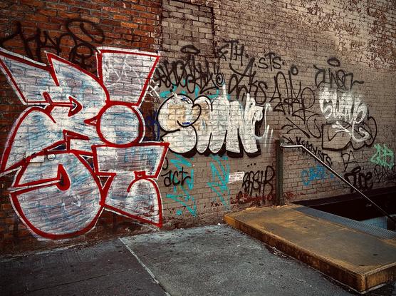 A color, wide-angle photograph that captures a weathered brick wall that serves as an urban canvas, heavily layered with various styles of graffiti next to a subway entrance in the Park Slope neighborhood of Brooklyn. On the left, a large, blocky wildstyle piece dominates the space, featuring white fill with light blue highlights and a thick, vibrant red outline. To its right, a rounded throw-up is in white with a bold black outline, surrounded by smaller black and teal tags, scribbles, and handstyles that overlap across the fading brown and tan bricks. The foreground shows a concrete sidewalk and a subway entrance leading underground. A set of concrete stairs is partially visible, protected by a simple metal handrail and a yellow-painted safety curb. The lighting is warm and directional, casting soft shadows that emphasize the gritty texture of the masonry and the peeling paint of the older tags.