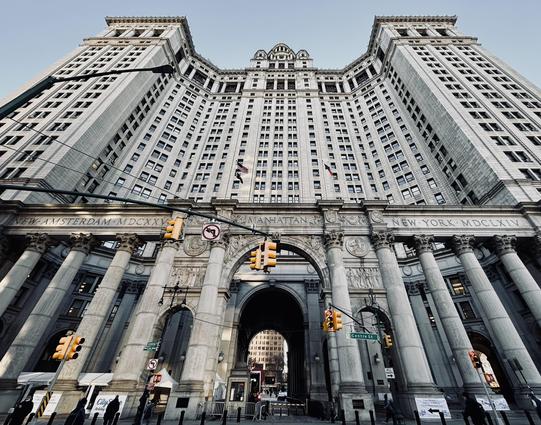 A color, wide-angle photograph in square aspect is ratio taken at a low angle that captures the imposing facade of the Manhattan Municipal Building in the Tribeca neighborhood of Manhattan. The architecture is a grand example of the Beaux-Arts style, featuring a massive central archway flanked by a series of soaring Corinthian columns. The building’s stone exterior is intricately carved with classical reliefs and the words "NEW AMSTERDAM MDCXXV," "MANHATTAN," and "NEW YORK MDCLXIV" inscribed across the frieze. Above the monumental base, the structure rises in a symmetrical, U-shaped configuration with hundreds of uniform windows, culminating in a tiered central tower. The foreground is busy with the traffic lights and sign elements for the intersection. Several bright yellow traffic signals hang from black wires that crisscross the frame, and a green street sign marks Centre Street. At the base of the columns, pedestrians walk past white temporary tents and security barriers. The lighting is crisp and clear, casting soft shadows that highlight the deep textures of the granite and the rhythmic patterns of the skyscraper’s many stories against a pale, cloudless winter morning sky.