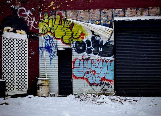 A color, wide-angle photograph in landscape orientation of the street scene along Fordham Road in the Bronx. A snow-covered lot sits in front of a weathered, industrial building featuring a mix of red brick and white siding. A large black metal roll-up door dominates the right side of the frame, while a smaller, narrow black door is centered against the white siding. To the left, a white lattice screen is set into a red brick archway. A layer of fresh snow blankets the ground, partially burying a weathered white barrel and a pile of bare, tangled branches in the foreground. The facade is heavily decorated with vibrant, overlapping graffiti tags in shades of lime green, bright blue, black, and red. The lime green bubble letters at the top and the large blue-and-white tag below it provide a sharp contrast to the peeling paint and exposed brick sections of the structure. Patches of snow cling to the tops of the doors and the uneven edges of the building’s exterior. The overcast afternoon sky is evenly illuminating the scene.