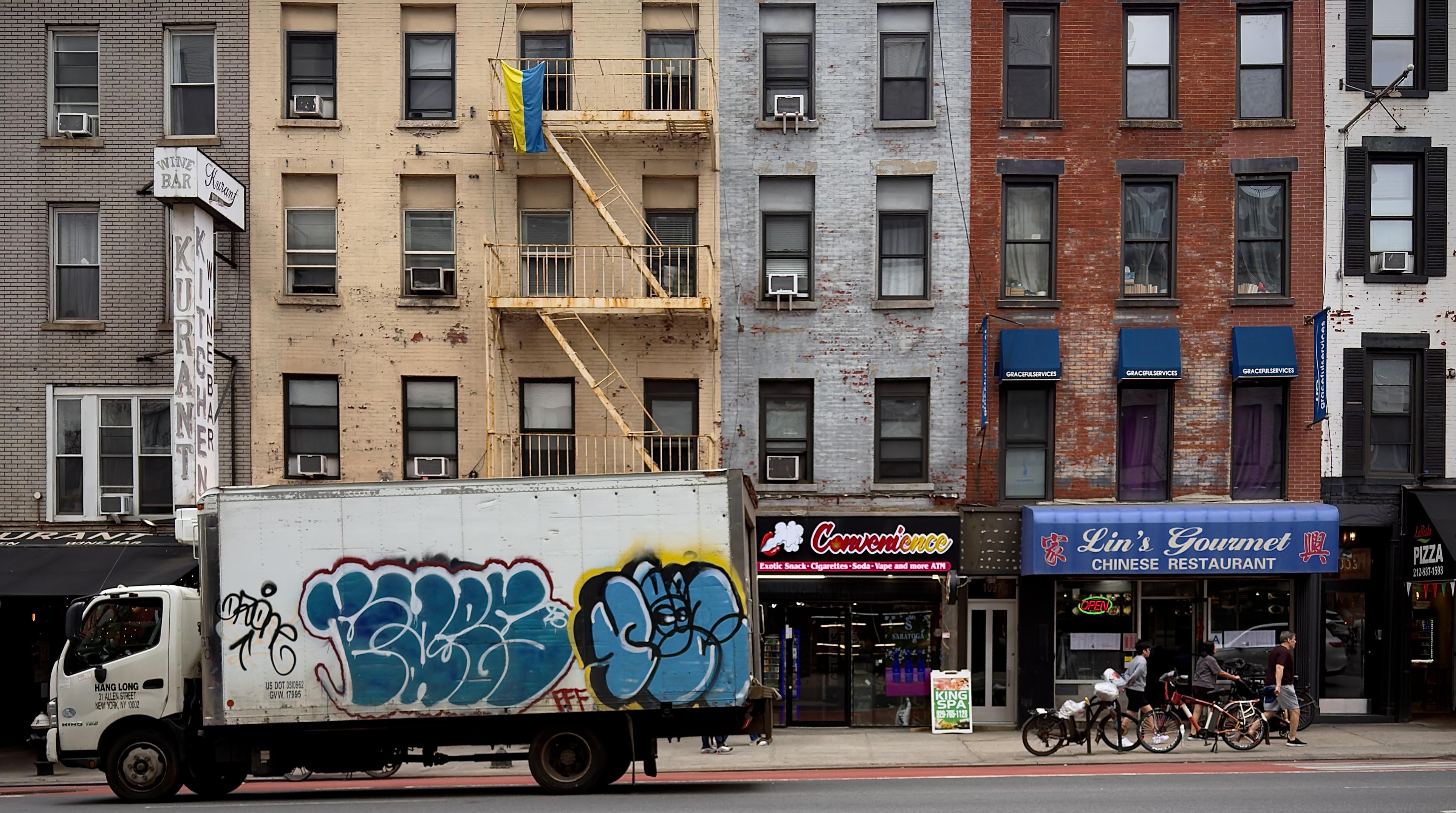 A color, wide-angle photograph in landscape orientation that captures a row of narrow, multi-story brick and masonry buildings, each featuring storefronts at the ground level. To the left, a tan building houses "Kurant Kitchen & Wine Bar" with its tall vertical sign, while the center and right buildings feature a convenience store and "Lin’s Gourmet Chinese Restaurant" with a prominent blue awning. A Ukrainian flag, with its distinct horizontal blue and yellow bands, hangs from a fire escape on the fourth floor of the tan building. On the sidewalk, several pedestrians are visible walking near a row of parked bicycles. In the foreground, a large white box truck is driving down the street towards the left side of the frame, partially obscuring the storefronts. The side of the truck is covered in bold, stylized graffiti. A large piece of "bubble-letter" graffiti dominates the center of the truck, featuring thick blue fill and bright yellow outlines that mirror the colors of the Ukrainian flag hanging directly above it on the building. The composition creates a striking visual parallel, tying the two elements together through their shared color palette against the backdrop of the weathered brick facades. The overcast afternoon sky is evenly illuminating the scene.