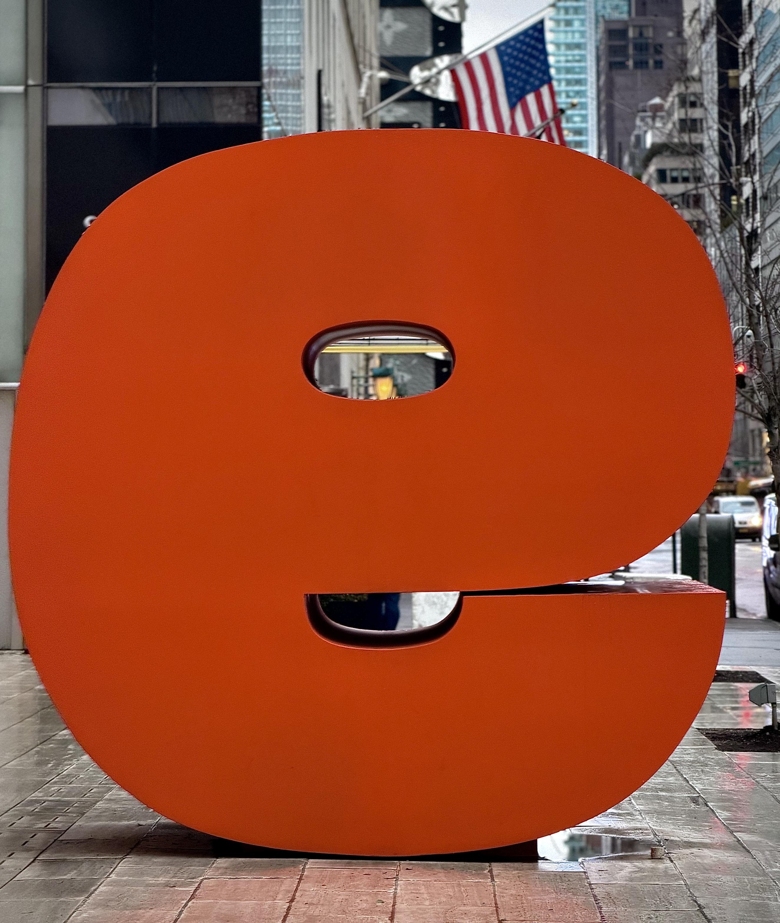 A color, medium-angle photograph that captures a massive, warm toned orange sculpture of a lowercase letter "e" positioned on a rain-slicked sidewalk along 57th Street in the Midtown East neighborhood of Manhattan on an overcast morning. The sculpture is rendered in a bold, sans-serif typeface with a thick, heavy weight, standing several feet tall. Its vibrant, matte orange finish provides a sharp, modern contrast against the muted, gray tones of the surrounding urban environment. The "e" is oriented so that the viewer looks directly at its side profile, with the two hollow counters acting as windows that frame the distant street view and a warm glowing light in the background. The setting is a high-end streetscape characterized by towering skyscrapers and storefronts. In the upper right background, an American flag hangs prominently from a building. The ground consists of wet, reflective paving stones that catch the hue of the sculpture and the surrounding city lights. The depth of field is shallow, keeping the giant letter in sharp focus while the city traffic, a green trash receptacle, and the architectural details of the block blur softly into the distance.