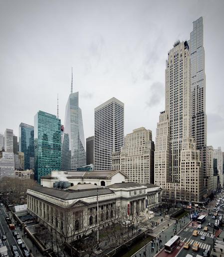 A color, high-angle photograph in portrait orientation that captures a bustling Manhattan streetscape on a gloomy, overcast day. The focal point is the Stephen A. Schwarzman Building (New York Public Library), a grand Beaux-Arts structure made of white marble. The library occupies the lower foreground along Fifth Avenue, featuring its iconic flat-topped roof, intricate cornices, and a grand staircase leading to Fifth Avenue. To its left, a small grove of bare, wintry trees lines the sidewalk. Wet pavement reflects the gray sky, and a few white plumes of steam rise from the building's rooftop vents, adding to the chilly, damp atmosphere of the scene. Surrounding the library is a dense collection of Midtown skyscrapers that stretch toward the top of the frame. To the left, the sleek, glass-clad Salesforce Tower and the pointed, crystalline spire of the Bank of America Tower dominate the skyline. On the right, the ultra-slender, tiered luxury residential towers of 520 Fifth Avenue and its neighbors rise vertically, showcasing a mix of modern glass and traditional masonry. Below, Fifth Avenue is active with city life; red and white transit buses, yellow taxis, and dark commuter cars navigate the rain-slicked asphalt, while tiny figures of pedestrians with umbrellas dot the sidewalks and crosswalks.