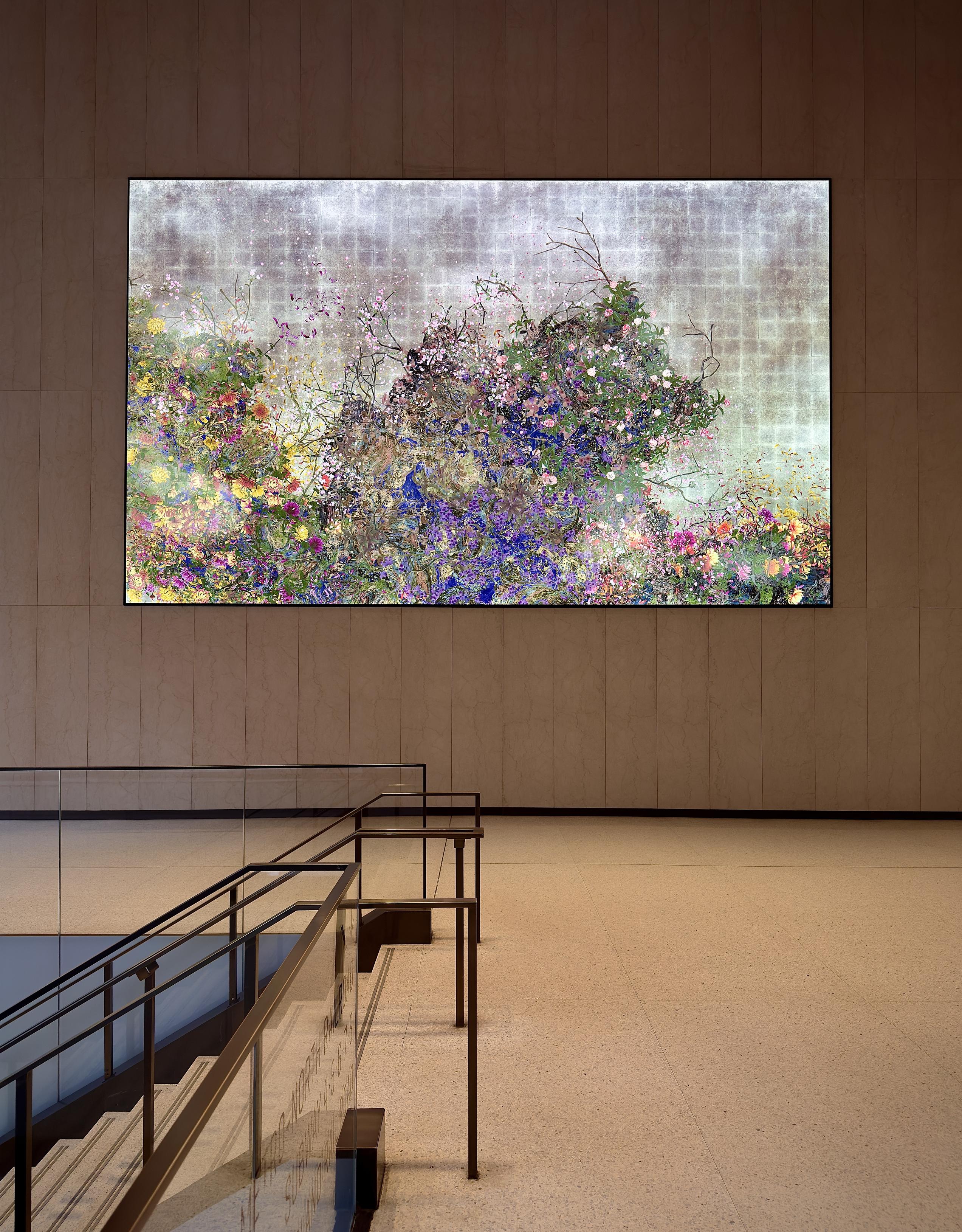 A color, wide-angle photograph in landscape orientation that captures the view of an interior landing within the One Vanderbilt building lobby in the Midtown East neighborhood of Manhattan. The composition is dominated by a gigantic, horizontal digital screen mounted on a minimalist, tan-colored stone wall. The screen displays a wildly vibrant floral animation characterized by a dense thicket of purple, pink, yellow, and blue flowers interwoven with green foliage and dark, spindly branches. The background of the digital art features a muted, textured gray and white grid pattern, creating a striking contrast between the organic, colorful chaos of the blossoms and the structured, industrial backdrop. In the foreground, the clean lines of the building’s modern architecture provide a sense of scale. A staircase with glass railings and sleek dark metal handrails descends into the lower left corner of the frame. The floor is a polished, light-colored terrazzo that reflects the soft, ambient glow from the screen and the overhead lighting. The vast, open space and the neutral tones of the stone walls emphasize the scale of the digital installation, making the floral animation appear as a window into a lush, synthetic garden. 