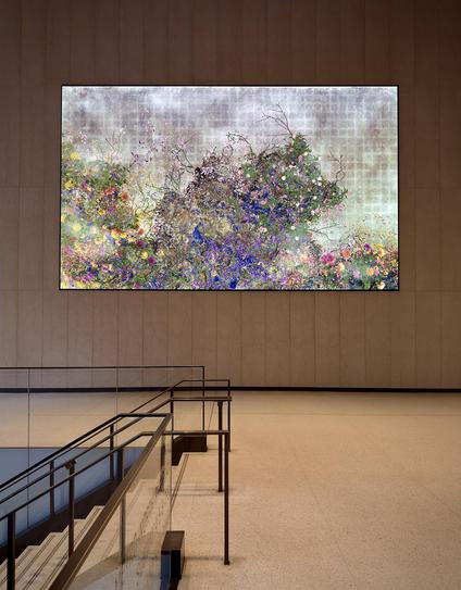 A color, wide-angle photograph in landscape orientation that captures the view of an interior landing within the One Vanderbilt building lobby in the Midtown East neighborhood of Manhattan. The composition is dominated by a gigantic, horizontal digital screen mounted on a minimalist, tan-colored stone wall. The screen displays a wildly vibrant floral animation characterized by a dense thicket of purple, pink, yellow, and blue flowers interwoven with green foliage and dark, spindly branches. The background of the digital art features a muted, textured gray and white grid pattern, creating a striking contrast between the organic, colorful chaos of the blossoms and the structured, industrial backdrop. In the foreground, the clean lines of the building’s modern architecture provide a sense of scale. A staircase with glass railings and sleek dark metal handrails descends into the lower left corner of the frame. The floor is a polished, light-colored terrazzo that reflects the soft, ambient glow from the screen and the overhead lighting. The vast, open space and the neutral tones of the stone walls emphasize the scale of the digital installation, making the floral animation appear as a window into a lush, synthetic garden. 