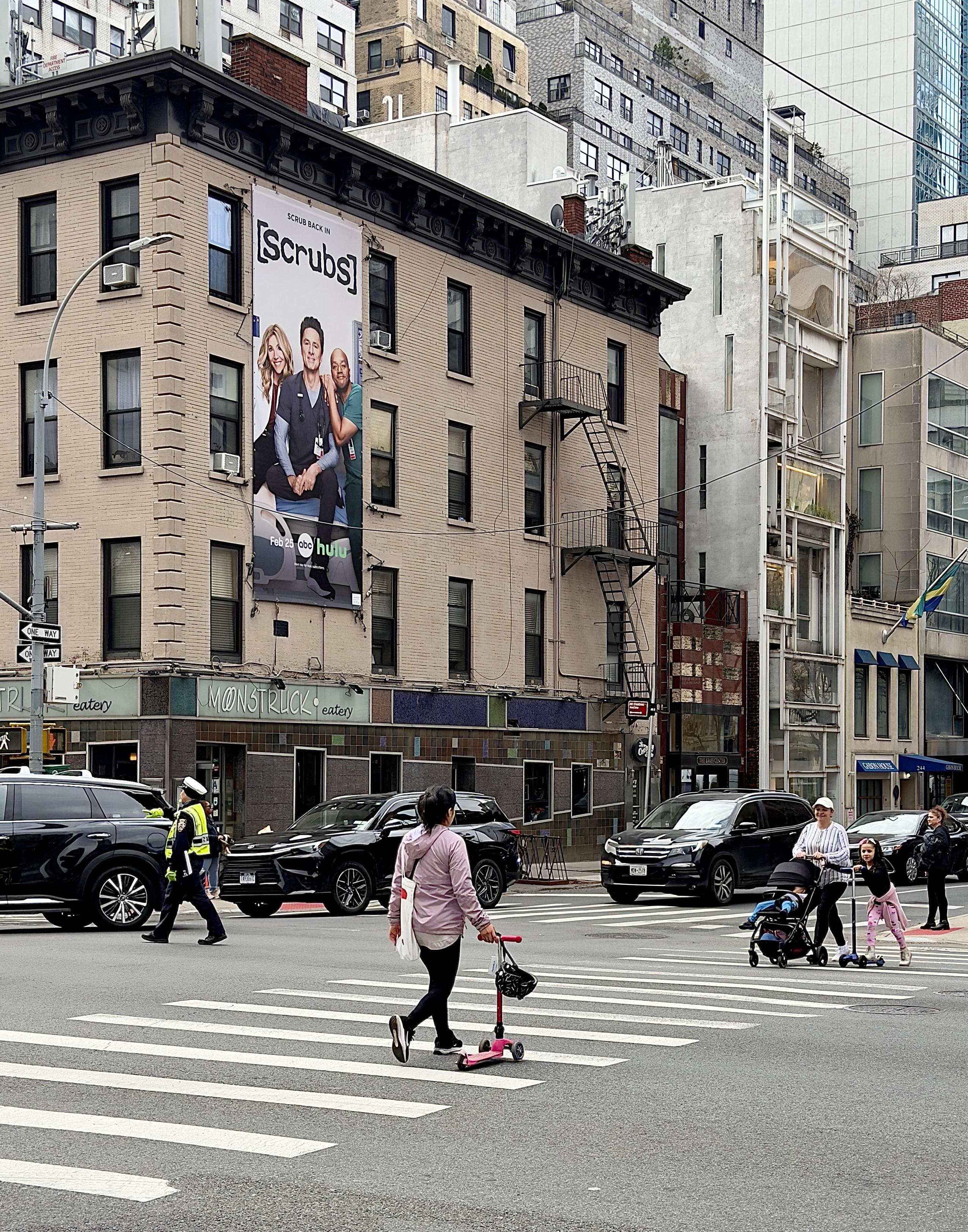 A color, wide-angle photograph in portrait orientation that captures the intersection of 2nd Avenue and East 58th Street, viewed from a pedestrian's perspective at the crosswalk. The focal point is a five-story, tan brick pre-war apartment building on the corner, which features a prominent vertical advertisement for a revival of the TV show Scrubs. The banner displays Sarah Chalke, Zach Braff, and Donald Faison sitting together with the show’s logo and a Hulu and ABC network credit at the bottom. Several dark SUVs are navigating the intersection, and a traffic officer in a neon yellow safety vest and white cap is visible directing the flow of vehicles. In the foreground, pedestrians are captured mid-stride across the wide, white-striped zebra crosswalk. A woman in a lilac hoodie and black leggings walks toward the left, pulling a small pink kick scooter behind her. To the right, a woman pushes a black stroller while a young girl on a blue scooter glides alongside her. The background showcases the dense architectural of the Midtown East neighborhood. The sky is overcast, casting a soft, even light over the scene.