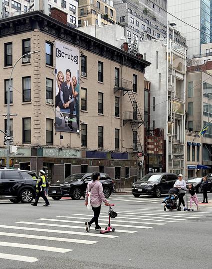 A color, wide-angle photograph in portrait orientation that captures the intersection of 2nd Avenue and East 58th Street, viewed from a pedestrian's perspective at the crosswalk. The focal point is a five-story, tan brick pre-war apartment building on the corner, which features a prominent vertical advertisement for a revival of the TV show Scrubs. The banner displays Sarah Chalke, Zach Braff, and Donald Faison sitting together with the show’s logo and a Hulu and ABC network credit at the bottom. Several dark SUVs are navigating the intersection, and a traffic officer in a neon yellow safety vest and white cap is visible directing the flow of vehicles. In the foreground, pedestrians are captured mid-stride across the wide, white-striped zebra crosswalk. A woman in a lilac hoodie and black leggings walks toward the left, pulling a small pink kick scooter behind her. To the right, a woman pushes a black stroller while a young girl on a blue scooter glides alongside her. The background showcases the dense architectural of the Midtown East neighborhood. The sky is overcast, casting a soft, even light over the scene.
