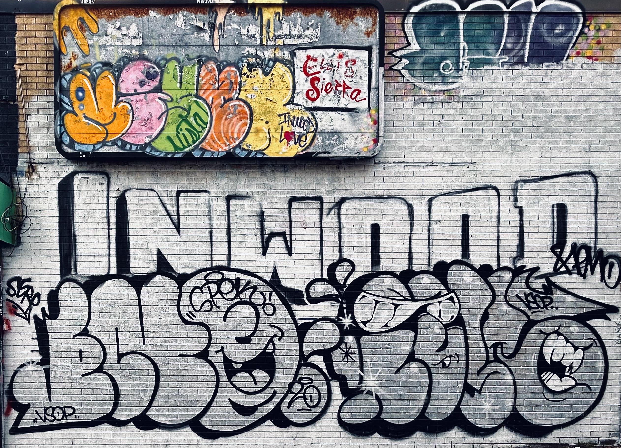 A color, wide-angle photograph in landscape orientation that captures a textured tan brick wall heavily decorated with various styles of graffiti. The majority of the wall has been covered in a base of white paint. At the center, the word "INWOOD" is rendered in large, blocky, white capital letters with thin black outlines. Directly below this, a series of large, rounded bubble letters in grayscale fill the lower half of the frame. These bubble characters are expressive, featuring cartoonish faces with wide, smiling mouths, sharp teeth, and eyes hidden behind stylized sunglasses. Smaller tags and marks like "VSOP," "SPEK," and "SEKO" are scattered around these larger pieces, adding to the layered, urban aesthetic. In the upper-left corner, a rectangular metal sign with a weathered, rusted frame sits mounted to the wall. The sign is covered in a more colorful style of graffiti, featuring rounded, overlapping shapes in bright shades of orange, pink, green, peach, and yellow. A prominent red tag reading "Elvis Sierra" is written in a fluid, cursive script on a white section of this sign, while the phrase "Inwood Love" is tucked neatly below it. To the right of the sign, on the brickwork, additional dark blue and purple tags complement the main display but have been partially covered in white base paint, creating a sense of dense, competing street art that covers nearly every inch of the visible surface. The scene is evenly illuminated by the overcast morning sky.