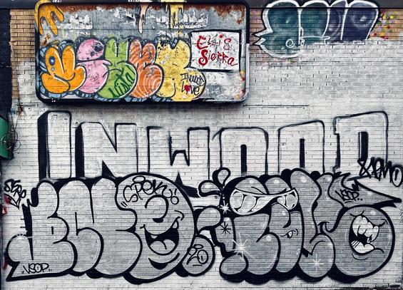 A color, wide-angle photograph in landscape orientation that captures a textured tan brick wall heavily decorated with various styles of graffiti. The majority of the wall has been covered in a base of white paint. At the center, the word "INWOOD" is rendered in large, blocky, white capital letters with thin black outlines. Directly below this, a series of large, rounded bubble letters in grayscale fill the lower half of the frame. These bubble characters are expressive, featuring cartoonish faces with wide, smiling mouths, sharp teeth, and eyes hidden behind stylized sunglasses. Smaller tags and marks like "VSOP," "SPEK," and "SEKO" are scattered around these larger pieces, adding to the layered, urban aesthetic. In the upper-left corner, a rectangular metal sign with a weathered, rusted frame sits mounted to the wall. The sign is covered in a more colorful style of graffiti, featuring rounded, overlapping shapes in bright shades of orange, pink, green, peach, and yellow. A prominent red tag reading "Elvis Sierra" is written in a fluid, cursive script on a white section of this sign, while the phrase "Inwood Love" is tucked neatly below it. To the right of the sign, on the brickwork, additional dark blue and purple tags complement the main display but have been partially covered in white base paint, creating a sense of dense, competing street art that covers nearly every inch of the visible surface. The scene is evenly illuminated by the overcast morning sky.