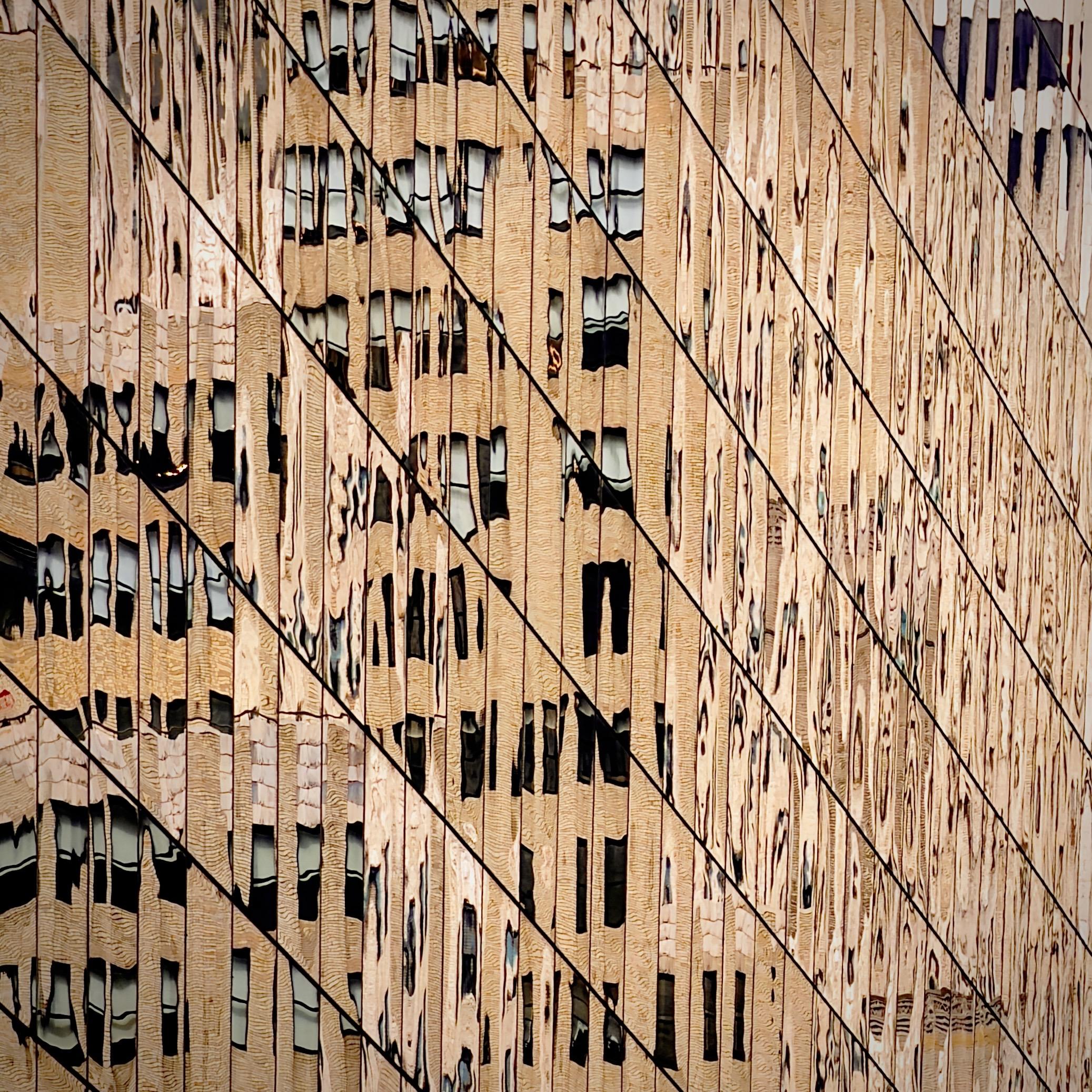 A color, medium-angle, close-up abstracted photograph of a Manhattan skyscraper featuring a grid of glass panels that reflect a neighboring stone building. The composition is dominated by a dizzying array of vertical and diagonal lines, with the thin, dark metallic frames of the glass panes creating a sharp geometric pattern across the frame. The reflected image is heavily distorted by the surface of the glass, causing the rows of windows from the across-the-street building to appear wavy, elongated, and liquid-like. The color palette is warm and monochromatic, consisting of rich tan, beige, and cream tones from the reflected masonry, contrasted by the dark, rhythmic lines of the window frames. The overall effect is one of architectural surrealism, where the rigid structure of the modern skyscraper dissolves into a fluid, shimmering golden mosaic. The perspective is tight and angled, focusing entirely on the play of light and shadow within the reflection rather than the structure of the building itself.